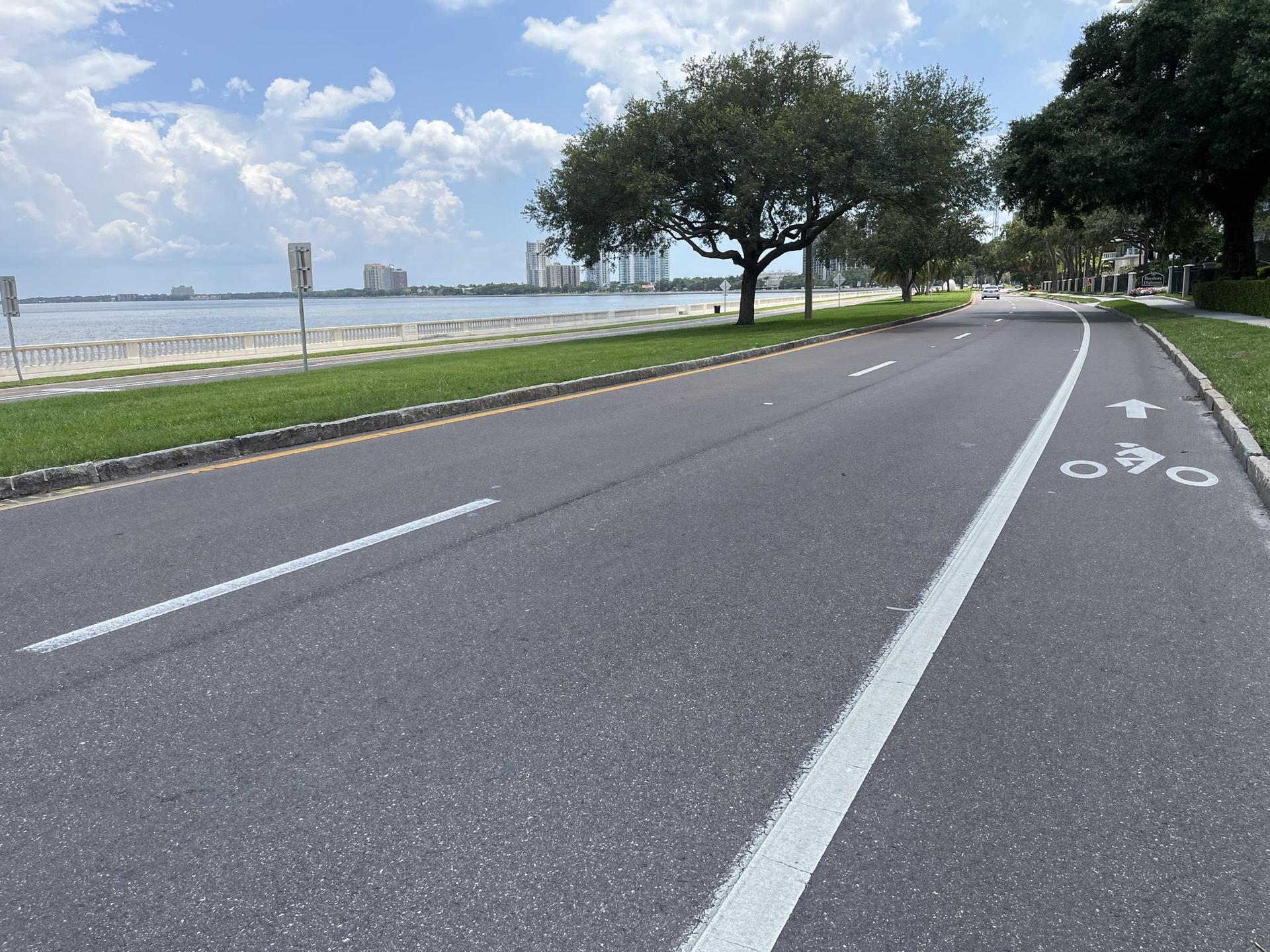 Coastal road with a bike lane, water, and distant buildings under a bright sky.