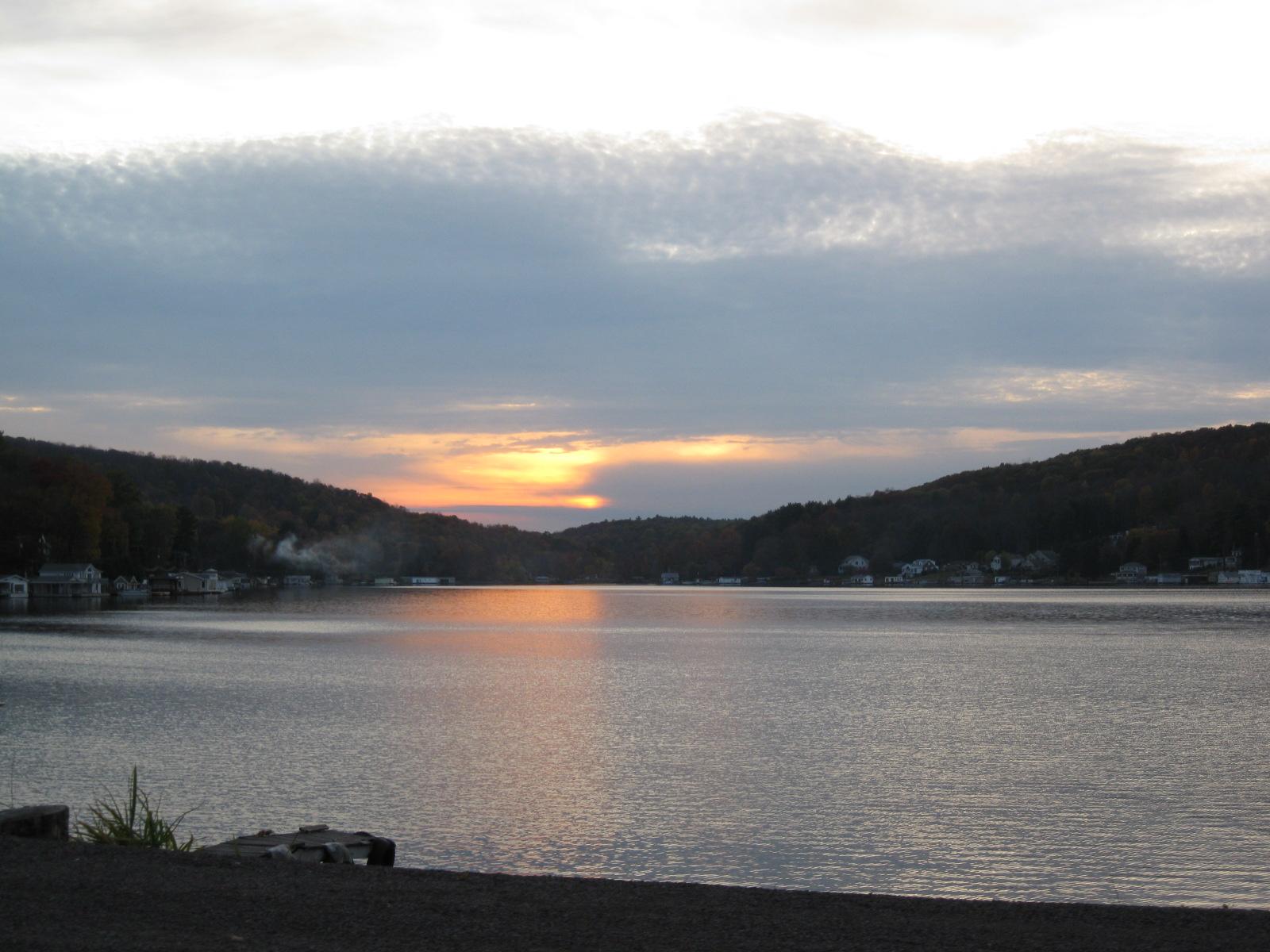 Orange sunset light reflects on a lake surrounded by wooded hills and distant houses.