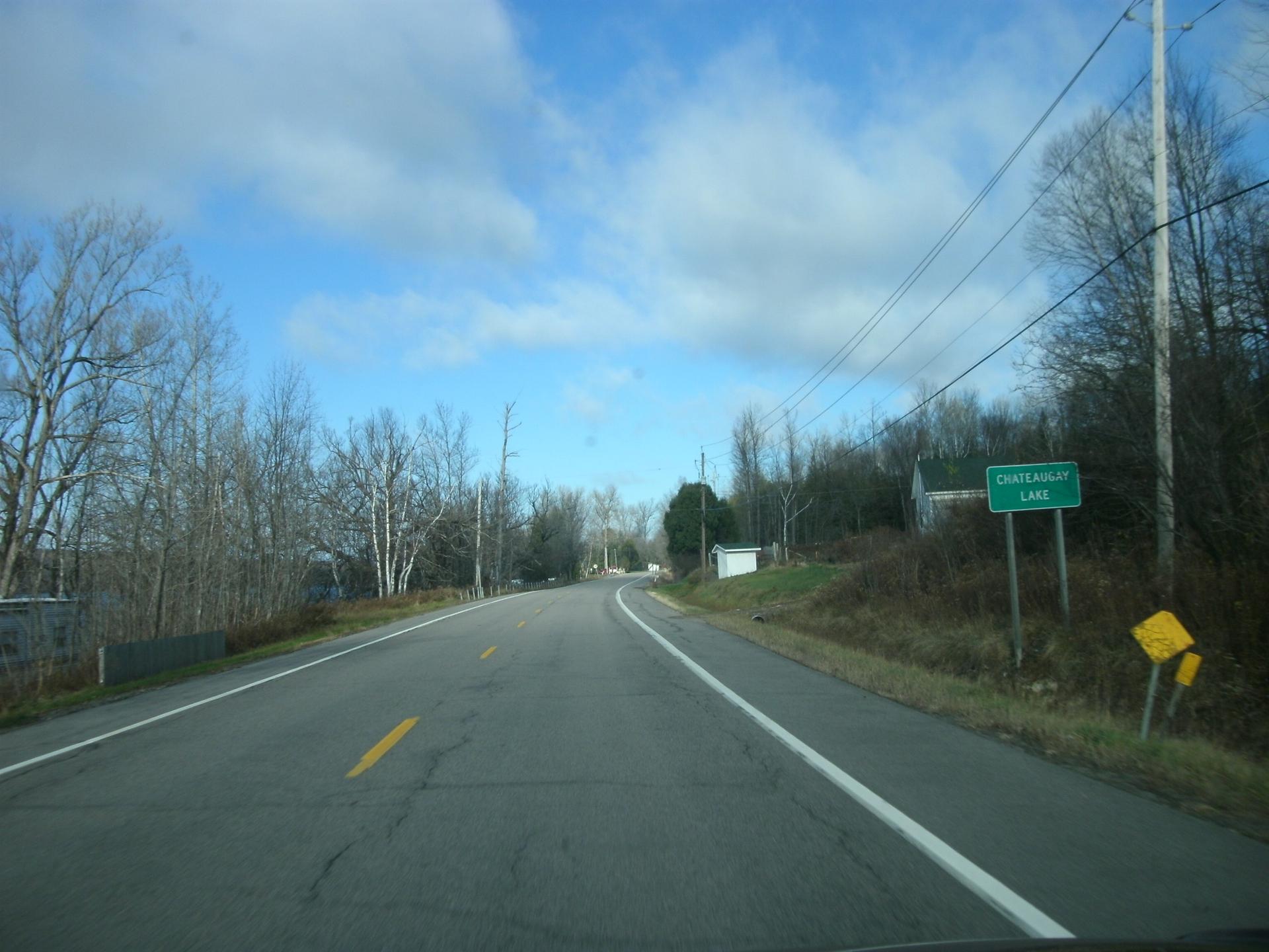 A paved road curves through a wooded landscape under a bright, cloudy sky.