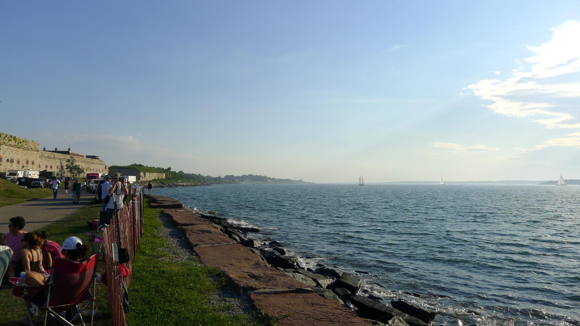 People relax along a rocky shore, watching distant boats on a sunny day.