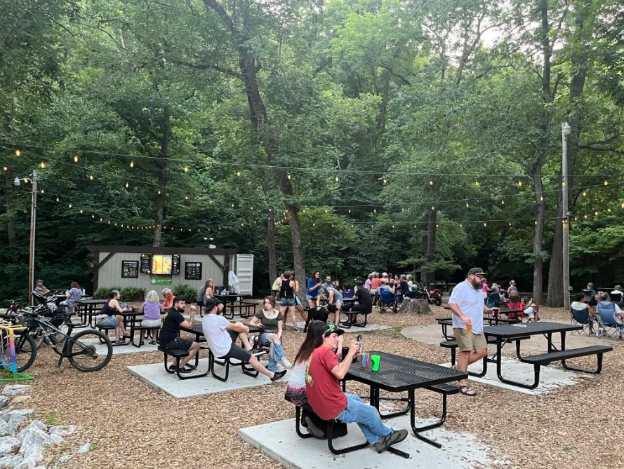 People socialize at picnic tables in a wooded outdoor area at dusk. - Green Bike Tower – Lake Bella Vista Trail loop from Bentonville