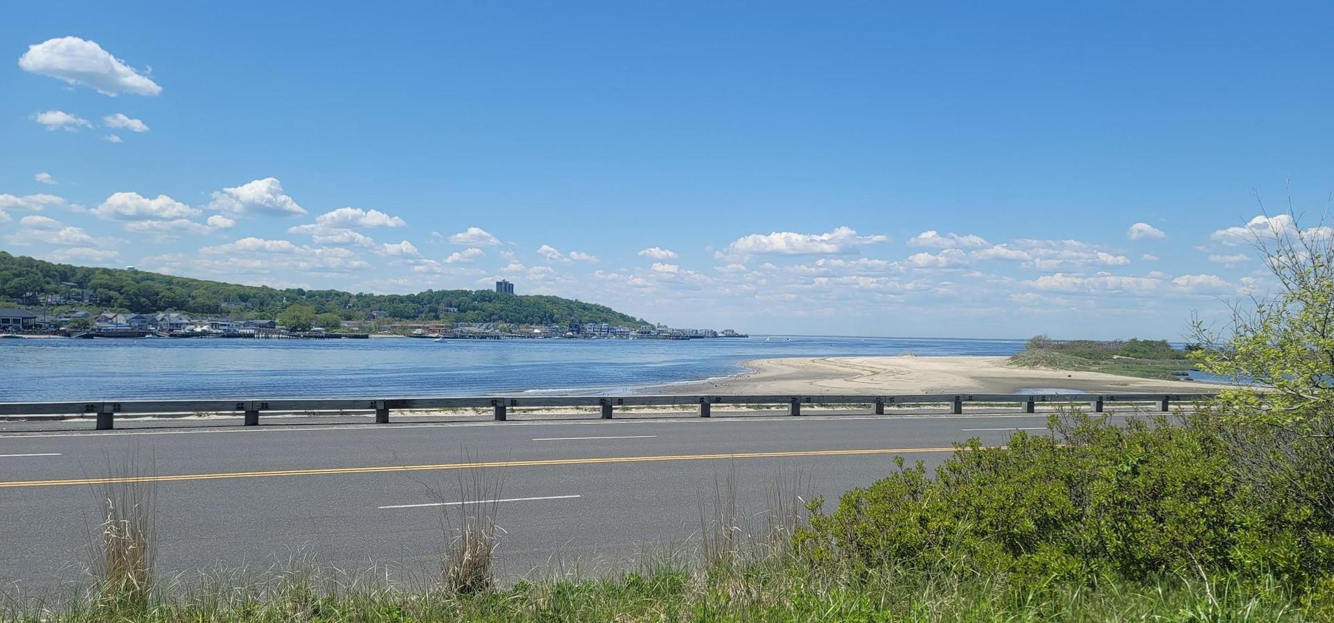 Coastal road next to calm water and a sandy beach on a sunny day.
