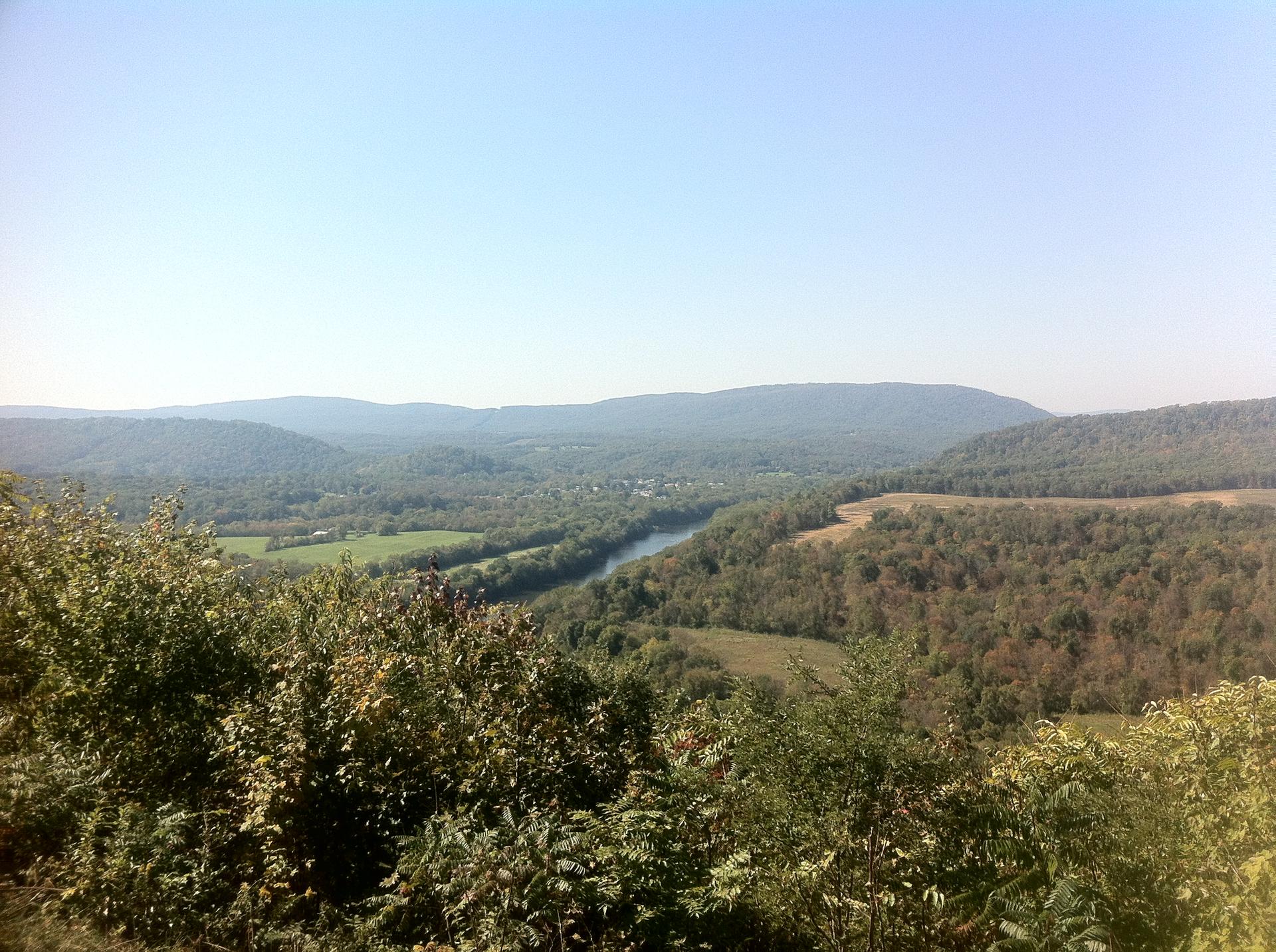 Bright daylight illuminates a winding river through a valley of forested hills.
