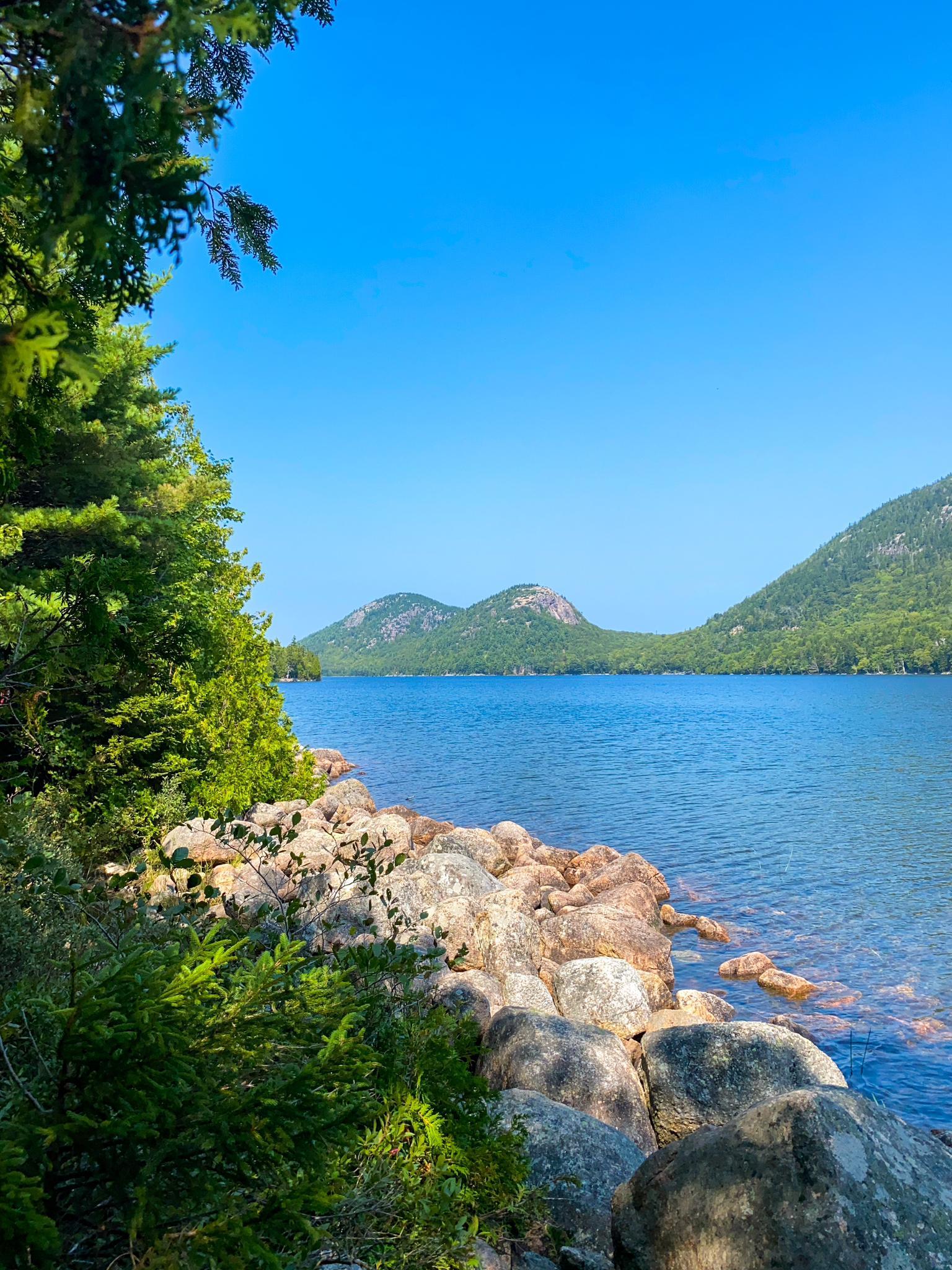 Clear blue lake with rocky shore, green trees, and distant mountains.