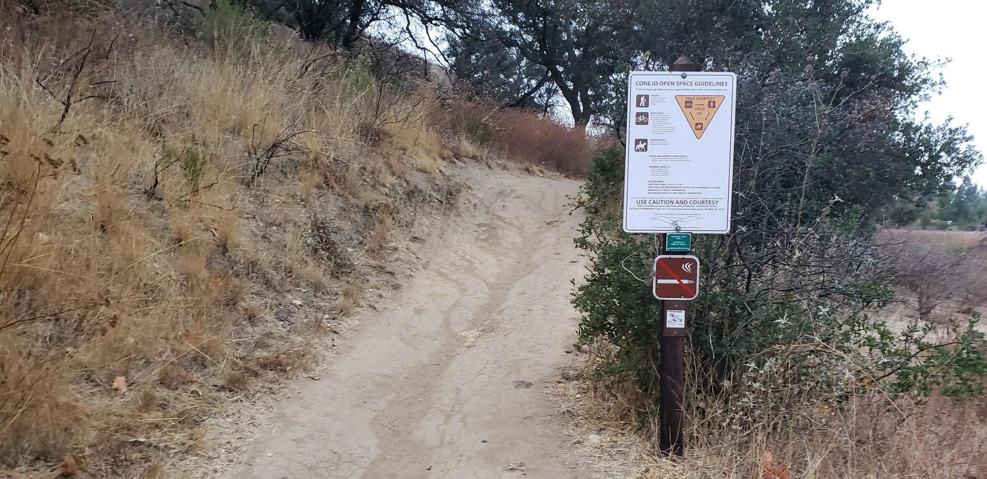 Dirt path leading uphill next to a trail sign and dry brush.