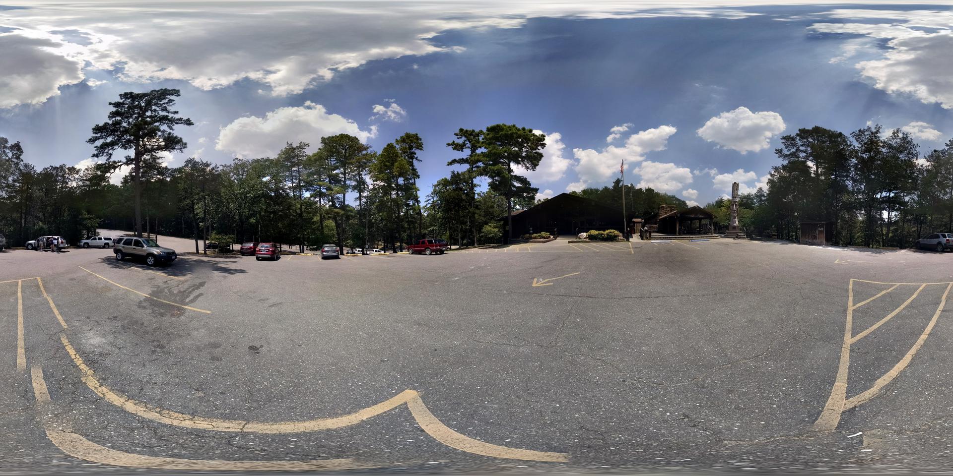 Bright parking lot with cars, tall trees, and a building under a partly cloudy sky.