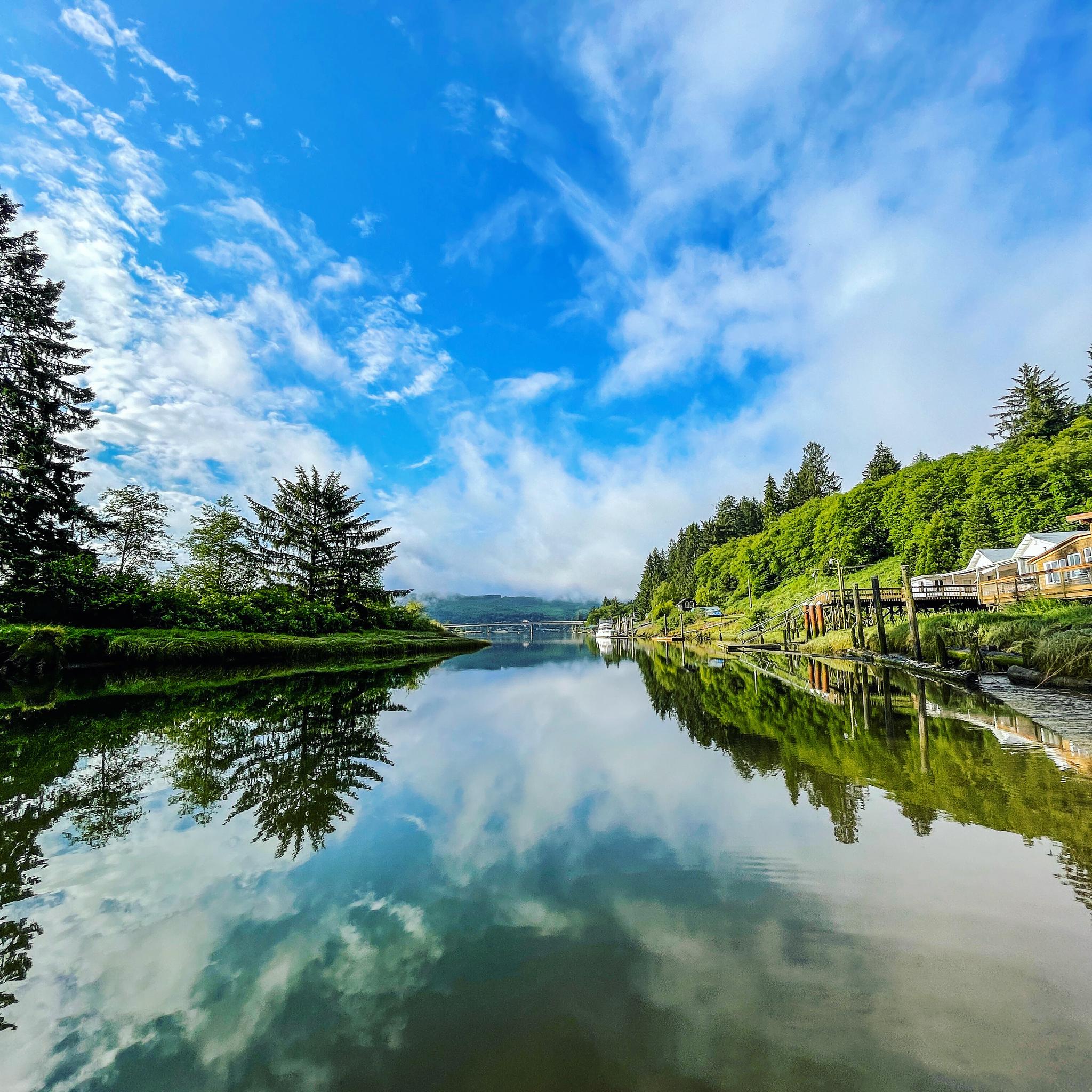 Calm river reflecting blue sky and clouds, with lush green banks and houses.
