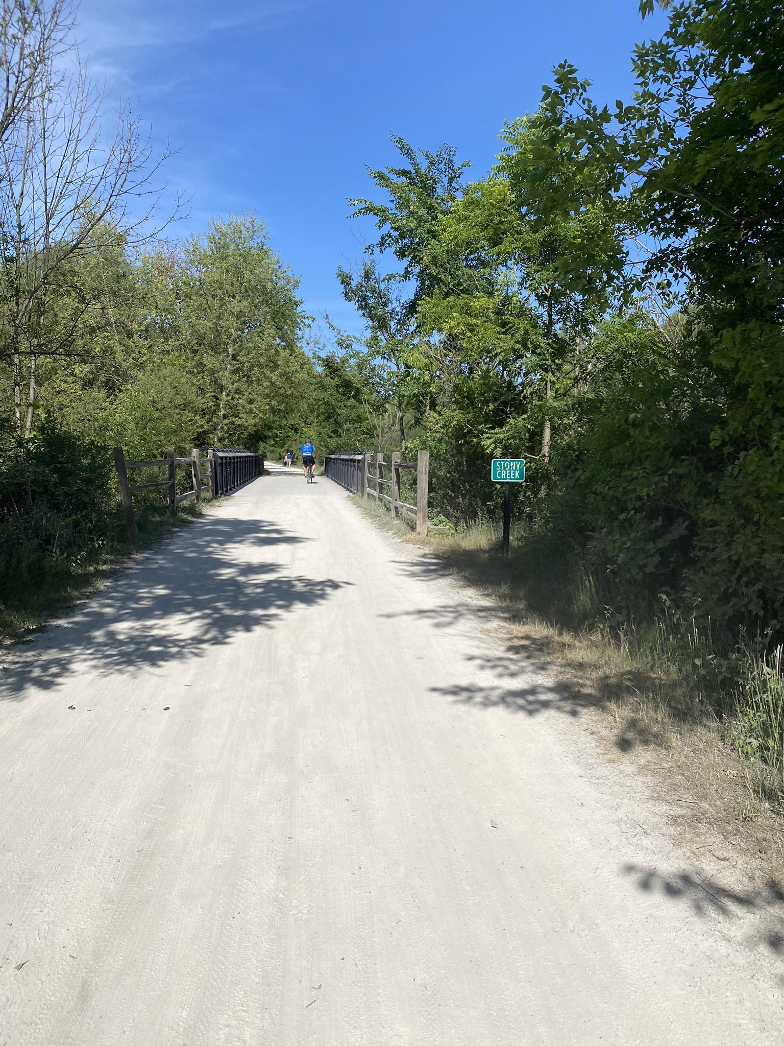 A person walks on a sunny gravel path through green trees.