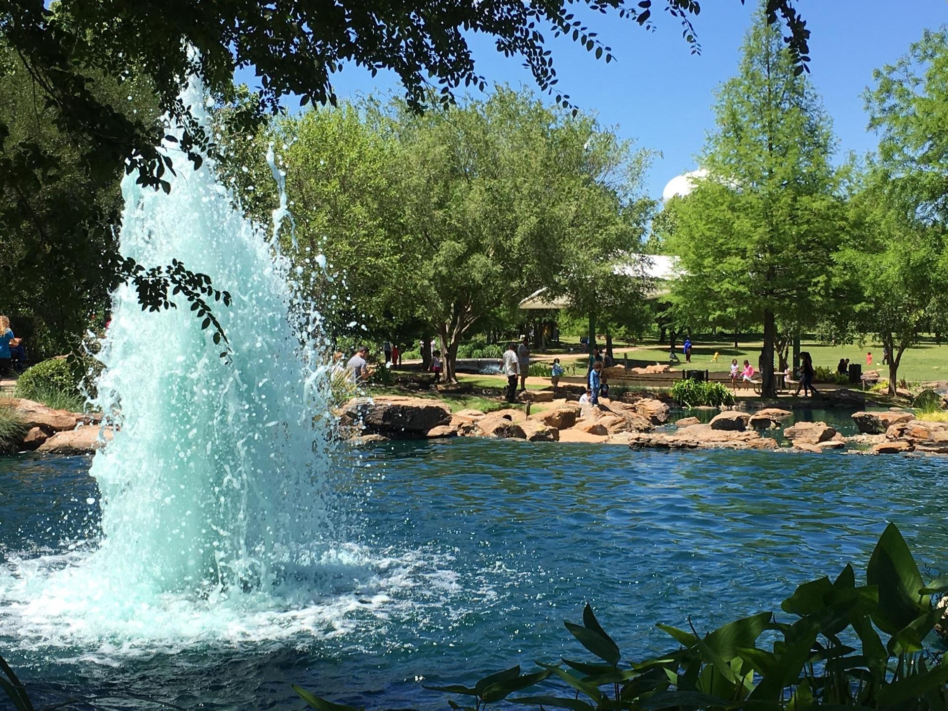 Bright fountain spraying water in a park pond with people and trees.