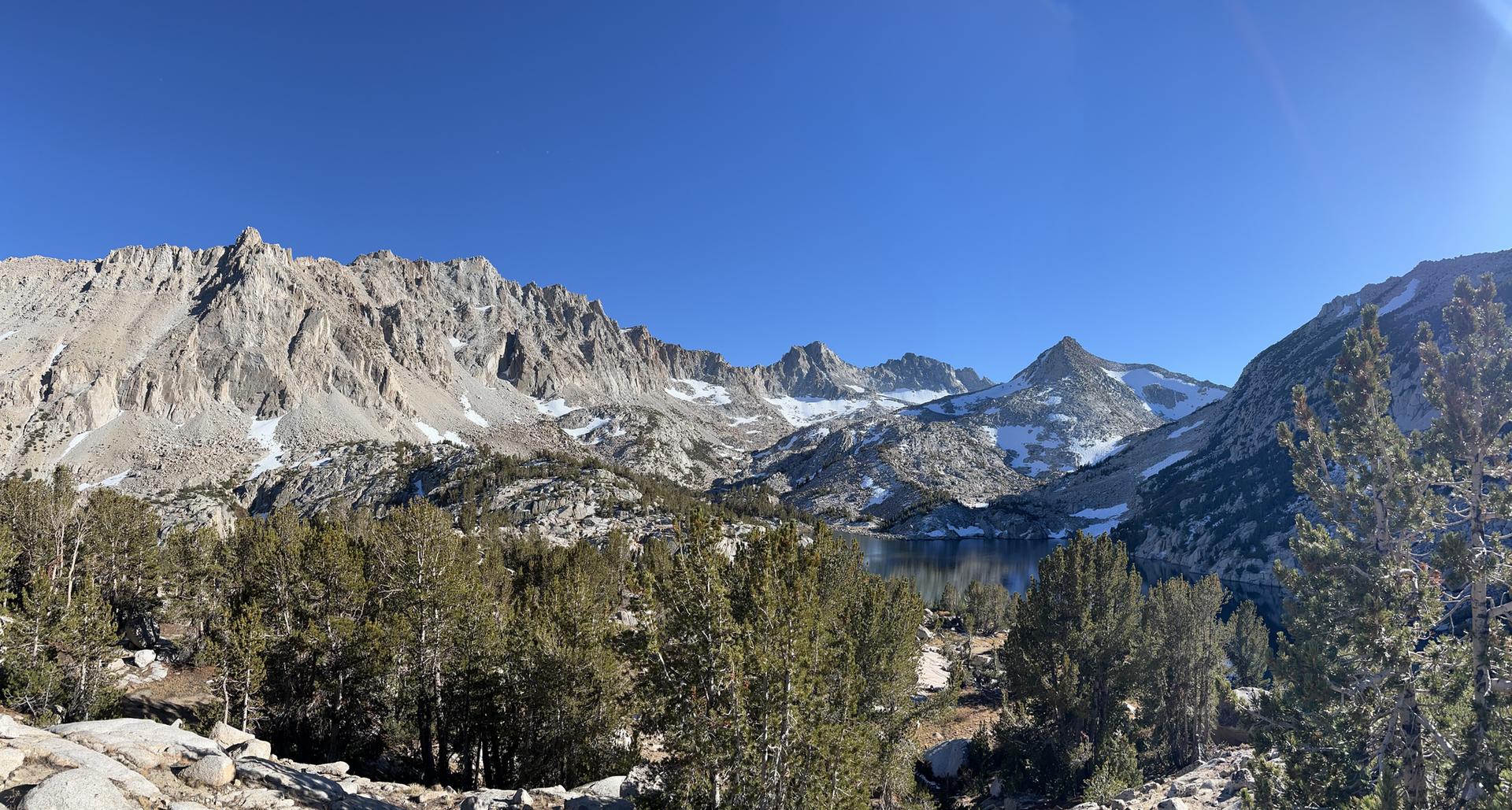 Bright panoramic view of rugged, snow-dusted mountains, a clear lake, and evergreen trees.