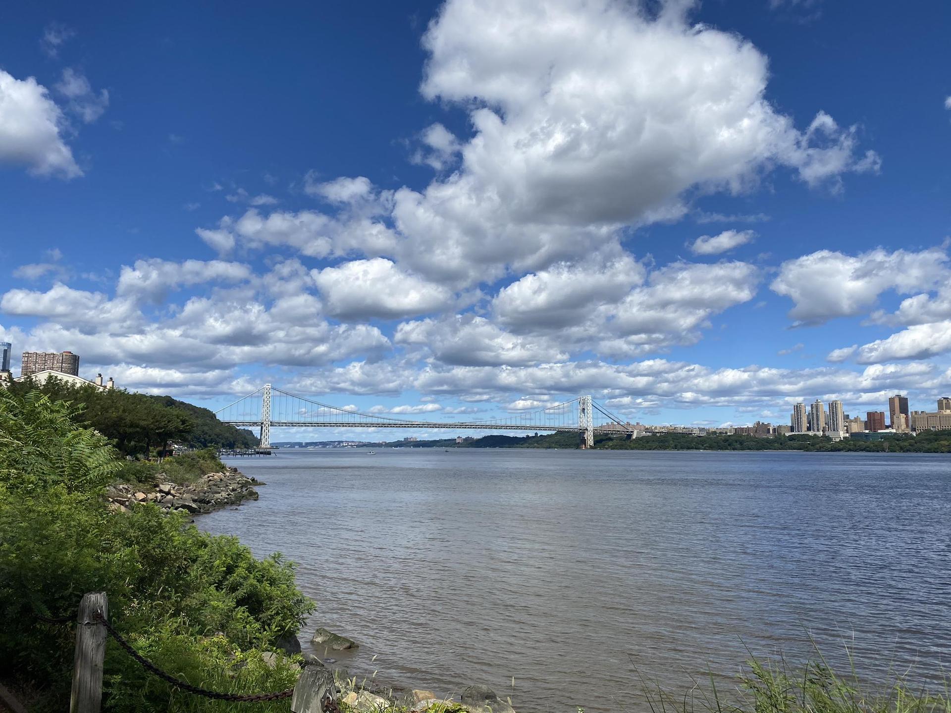 Wide river with a suspension bridge and distant city under a bright blue sky.
