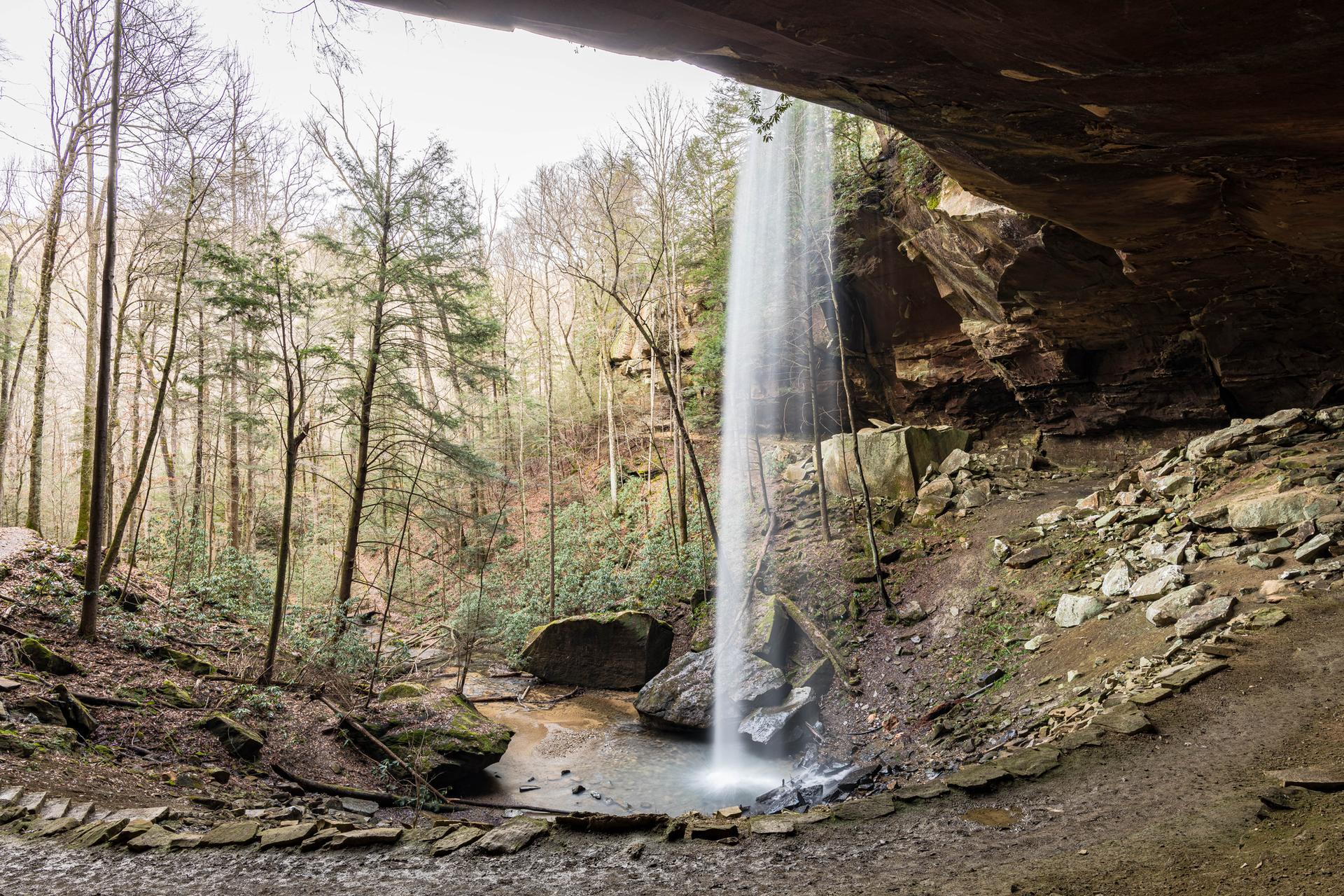 A tall waterfall plunges into a rocky pool under a large rock overhang.