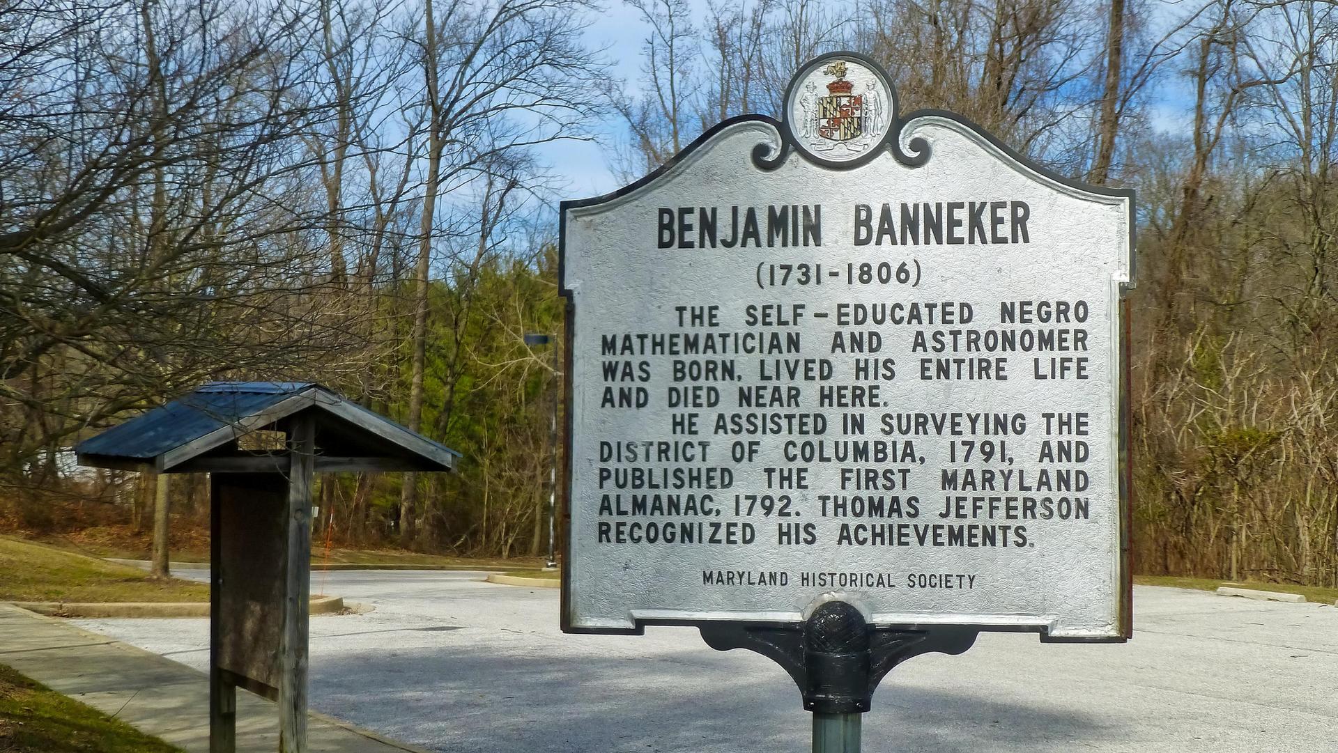 A historical marker sign and a smaller wooden sign stand among bare trees.