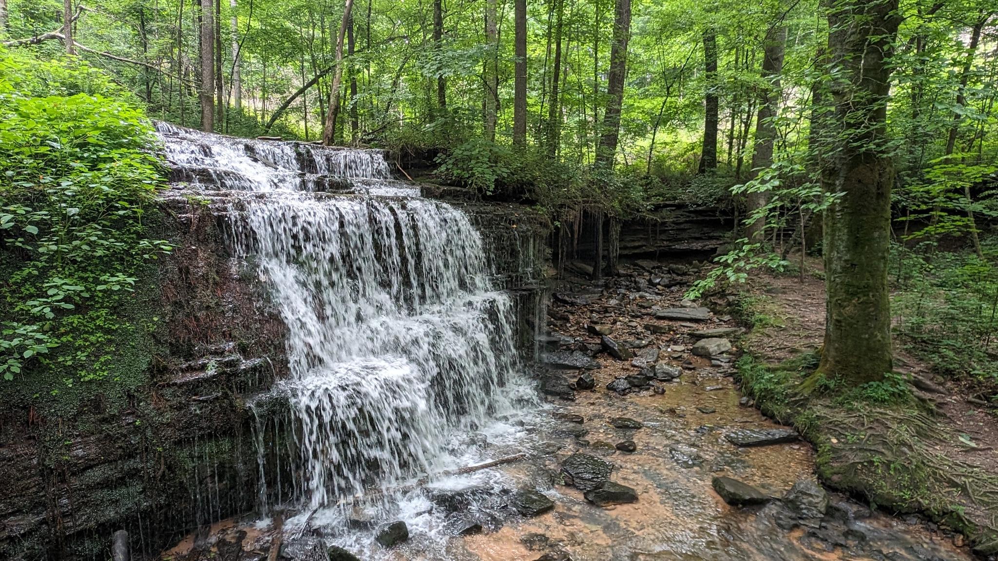 Multi-tiered waterfall cascades into a rocky stream in a lush green forest.