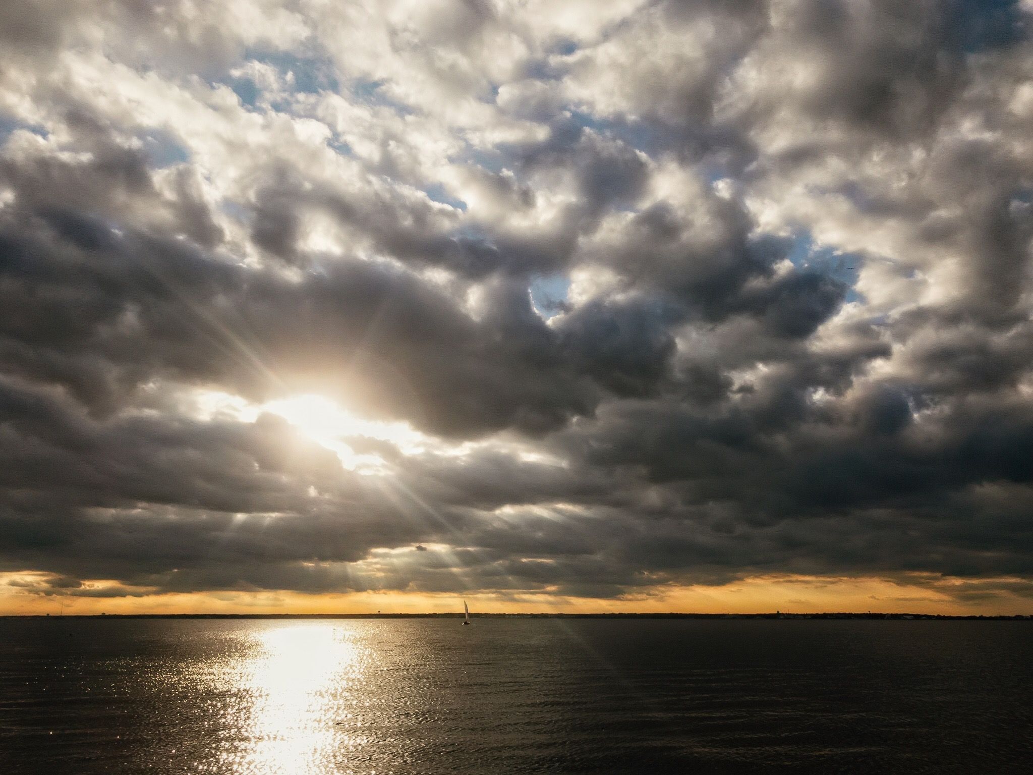 Sunbeams pierce dramatic clouds over a vast body of water with a distant sailboat. - Cattus Island County Park