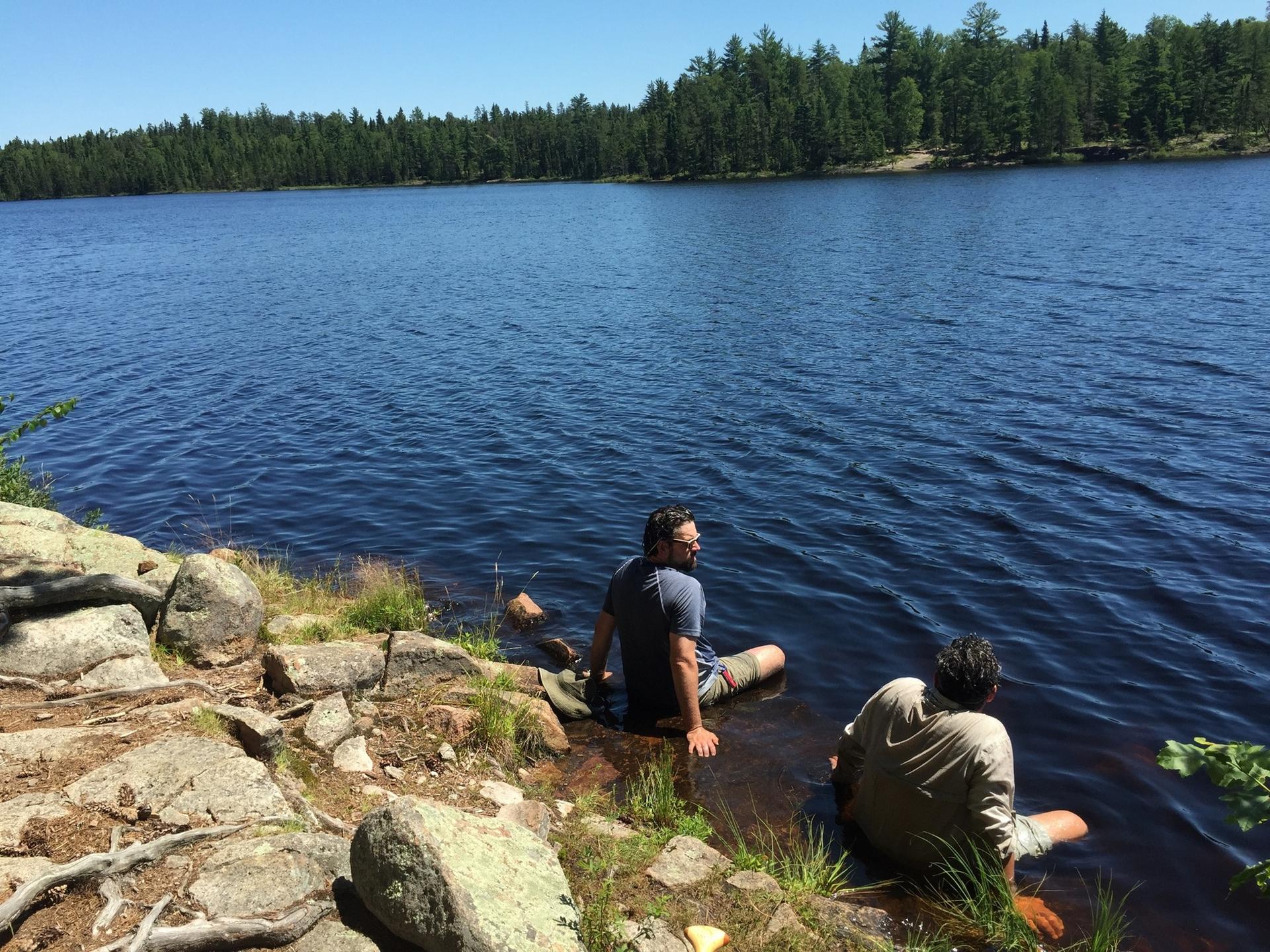 Two people relax on a rocky shore, feet in a sunny blue lake.