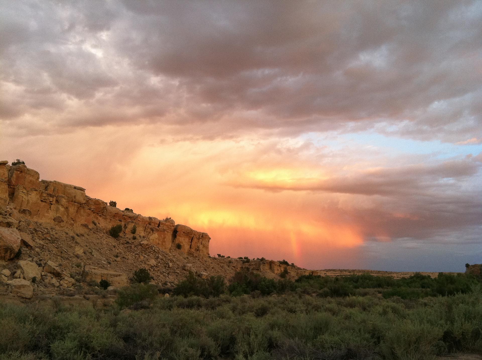Dramatic sunset sky with a rainbow over a desert cliff and brush.