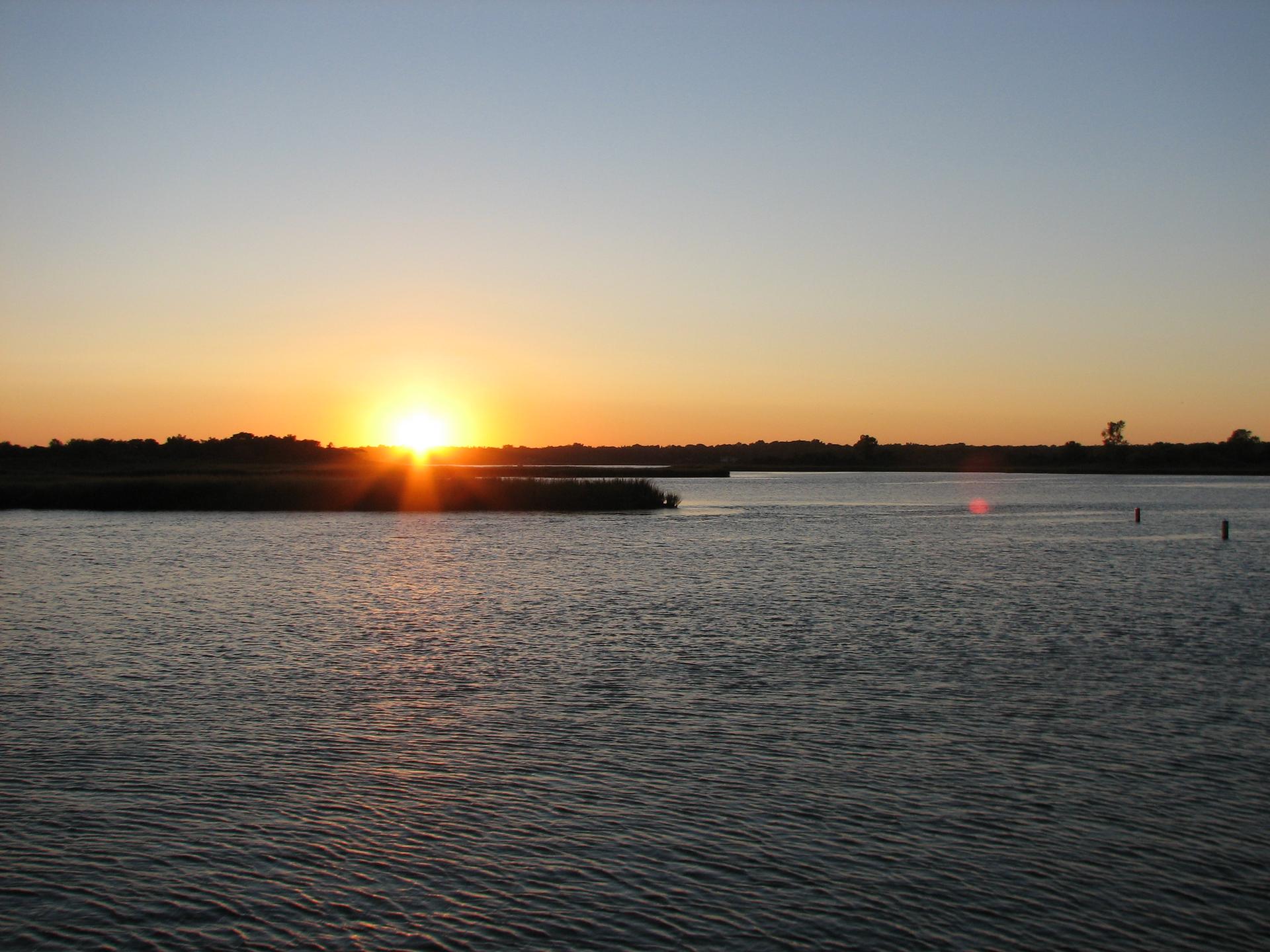 Golden sunset over calm water with distant marshland under a clear sky.