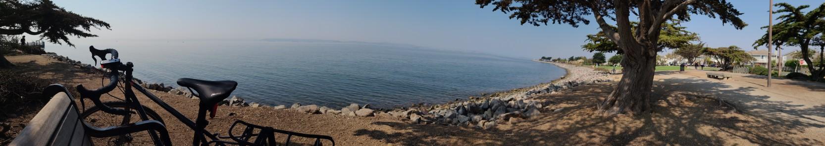 A panoramic view of a bicycle parked by a calm bay and sunny beach.