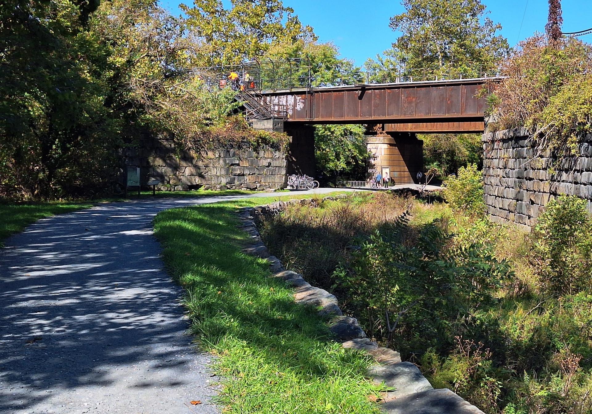 Gravel path passes under a rusty metal bridge with stone walls and green foliage.