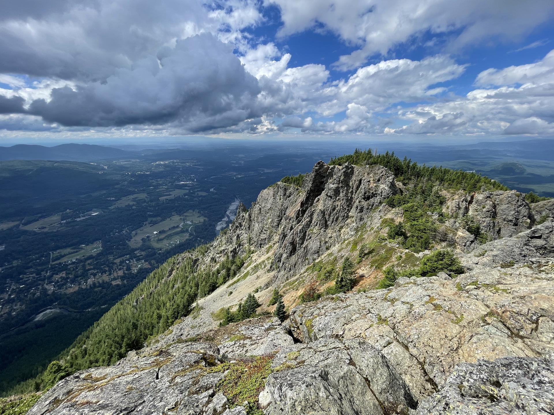 Rocky mountain ridge overlooks a vast forested valley under a cloudy sky.