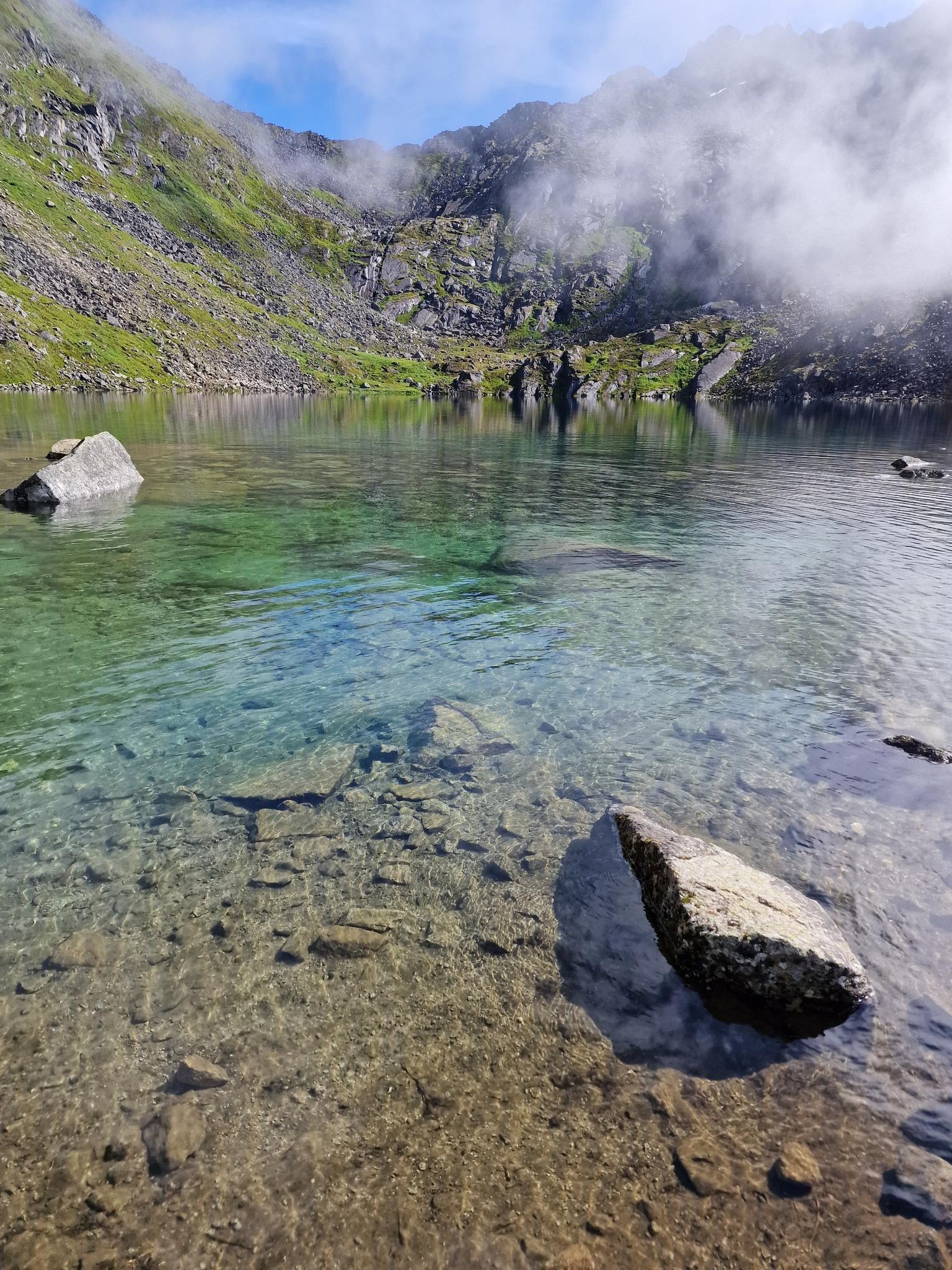 Clear turquoise mountain lake with rocky bottom, misty green slopes.