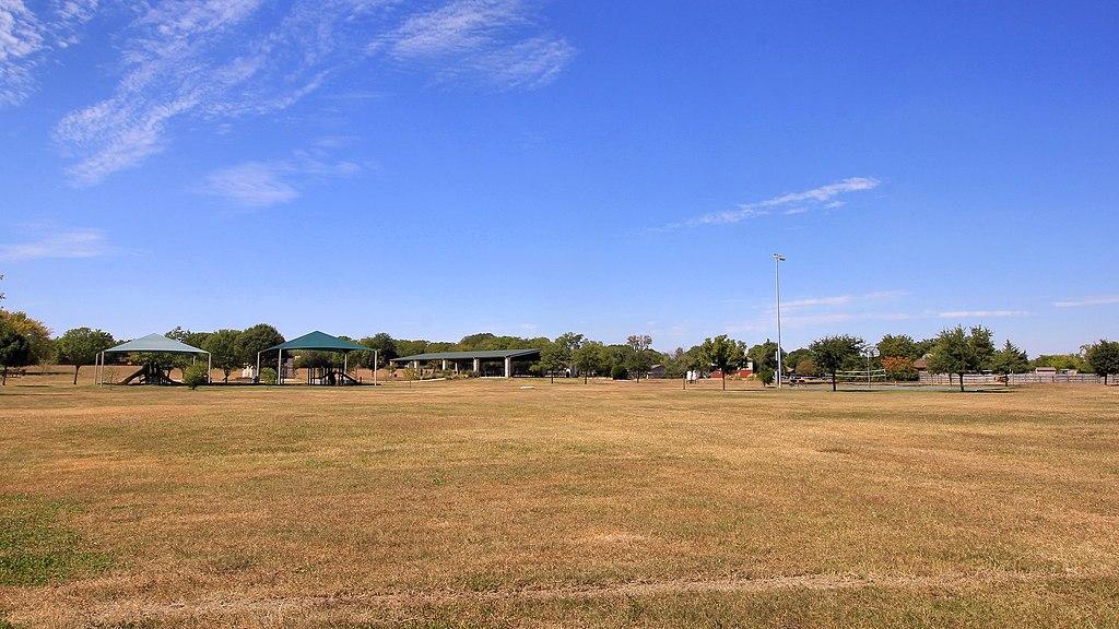 Expansive sunny grassy field with distant park structures under a blue sky.