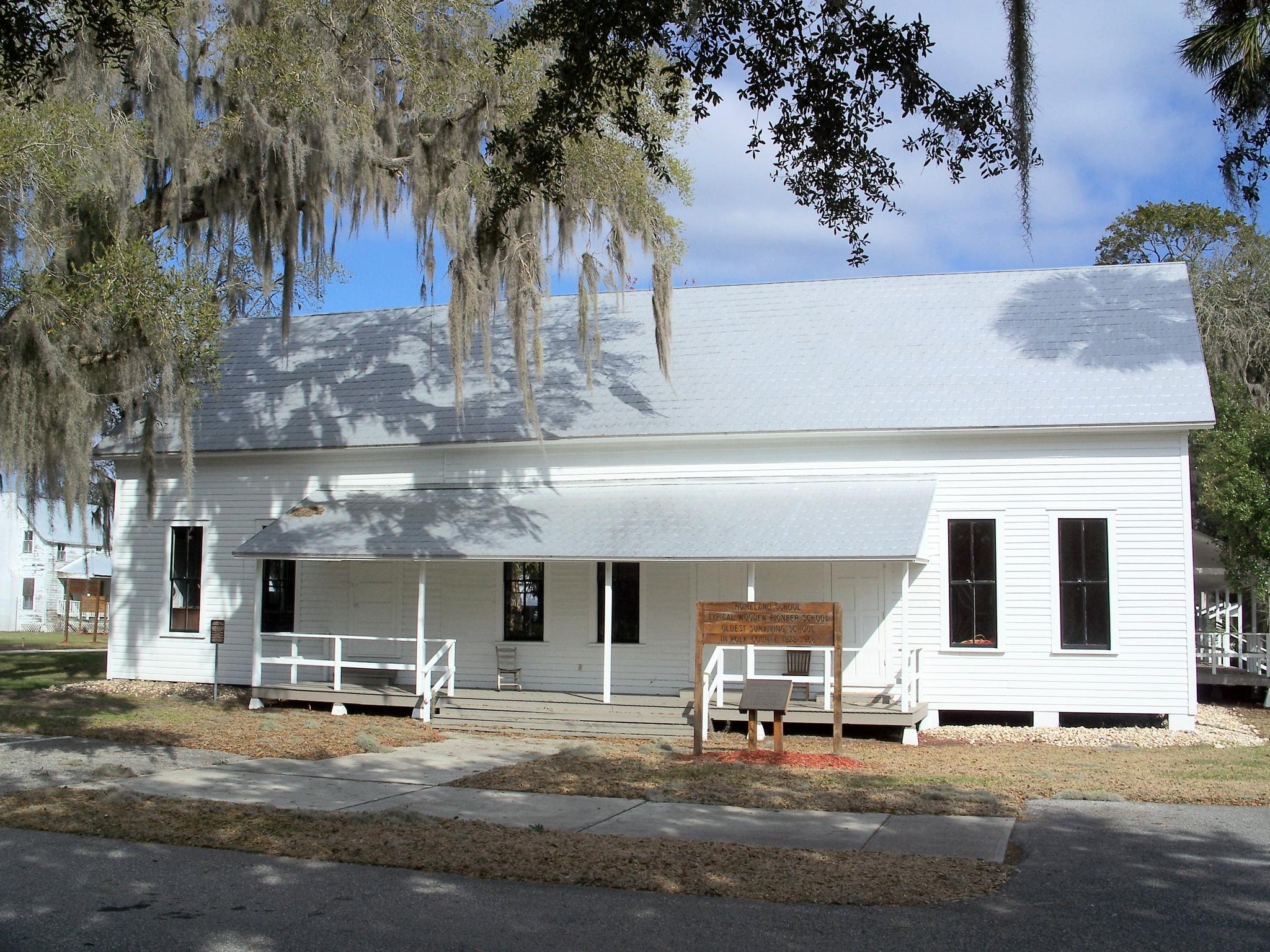 A white, old-fashioned building with a long porch, shaded by trees.