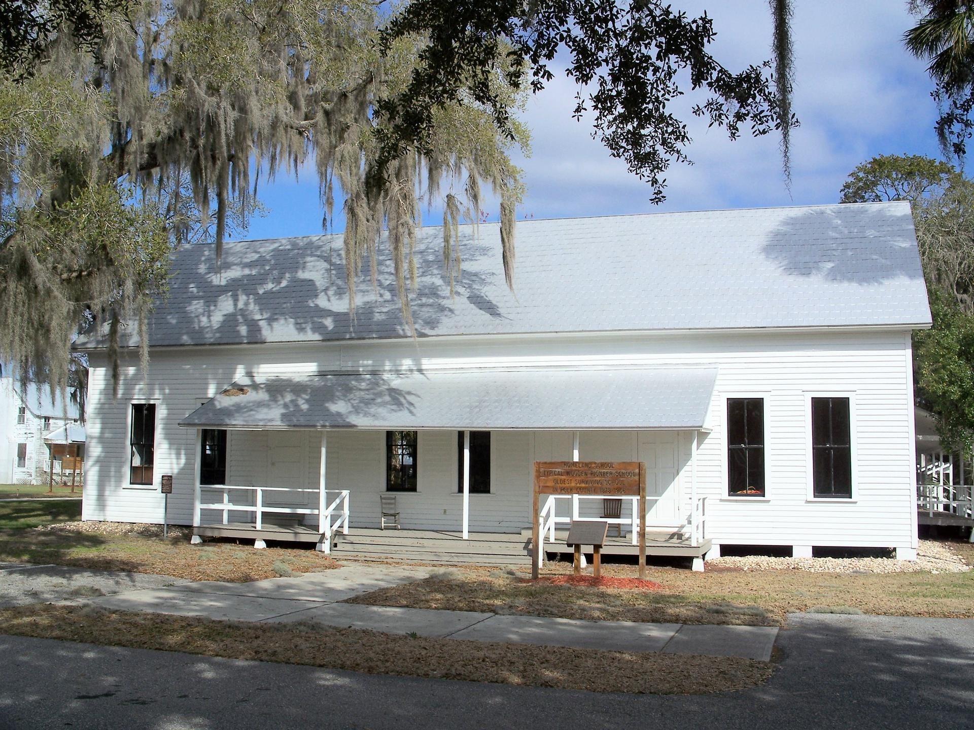 A white, old-fashioned building with a long porch, shaded by trees.