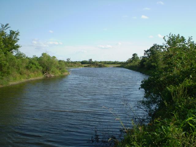 A wide, calm river flows between lush green banks under a blue sky.