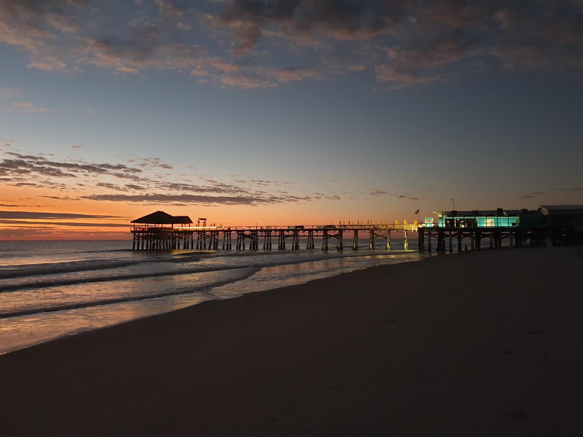 Long pier extends over ocean at sunset, with dark beach foreground.
