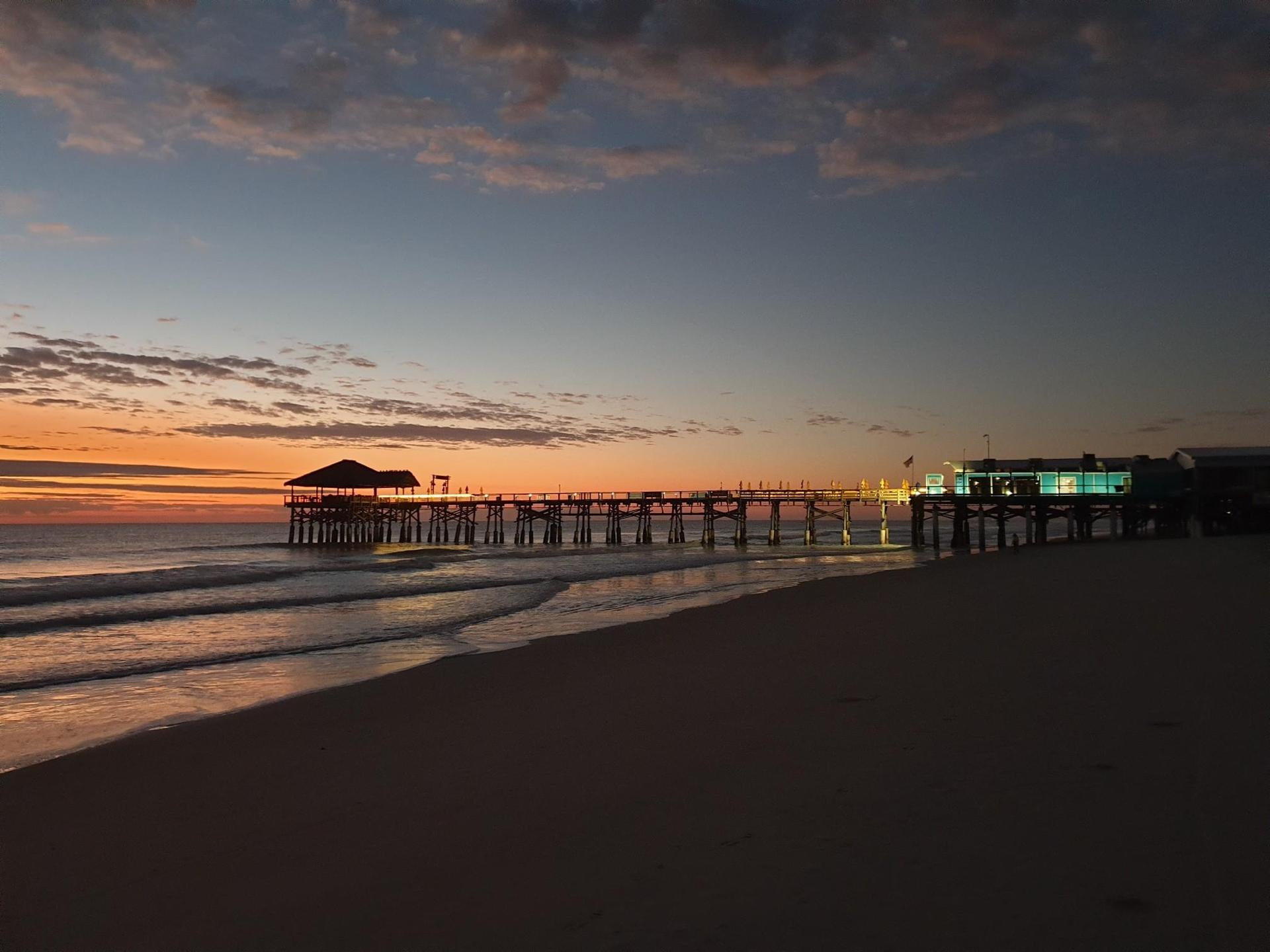 Long pier extends over ocean at sunset, with dark beach foreground.