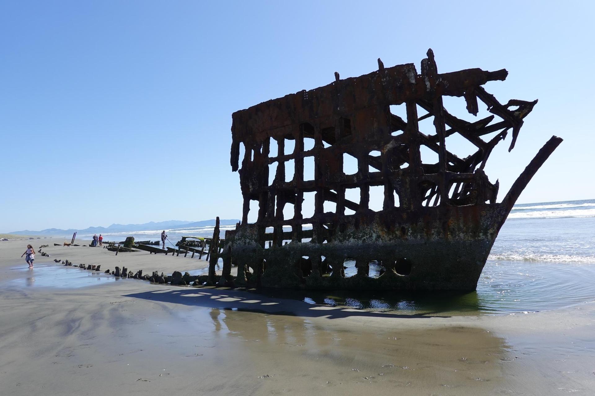 A large rusty shipwreck rests on a sunny sandy beach with people nearby.
