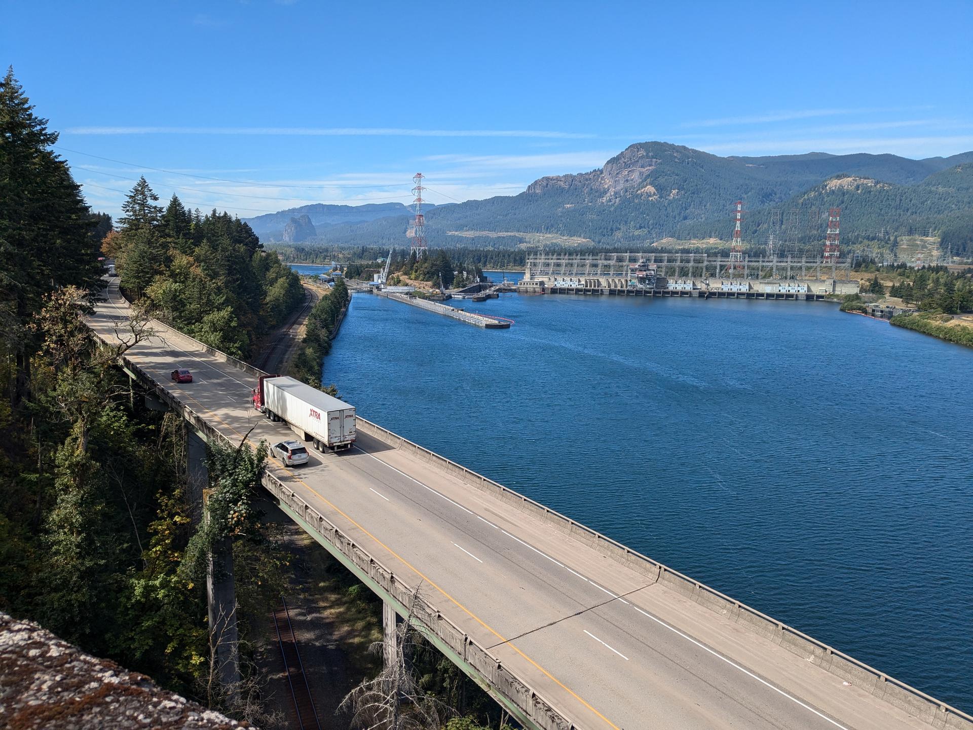 A truck crosses a bridge over a wide river with mountains and a dam.