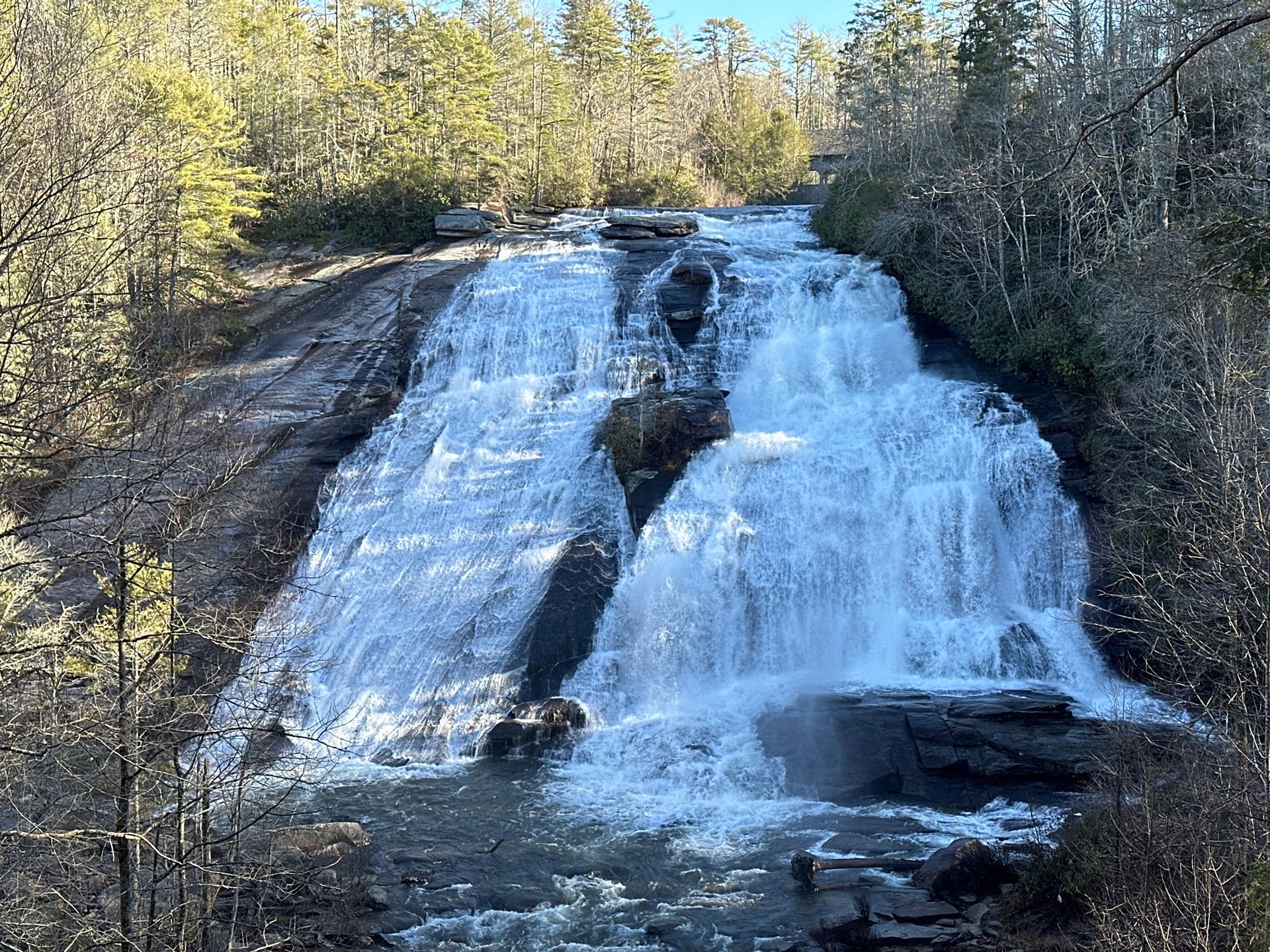 Large waterfall cascades over wide rock face into river on a sunny day.