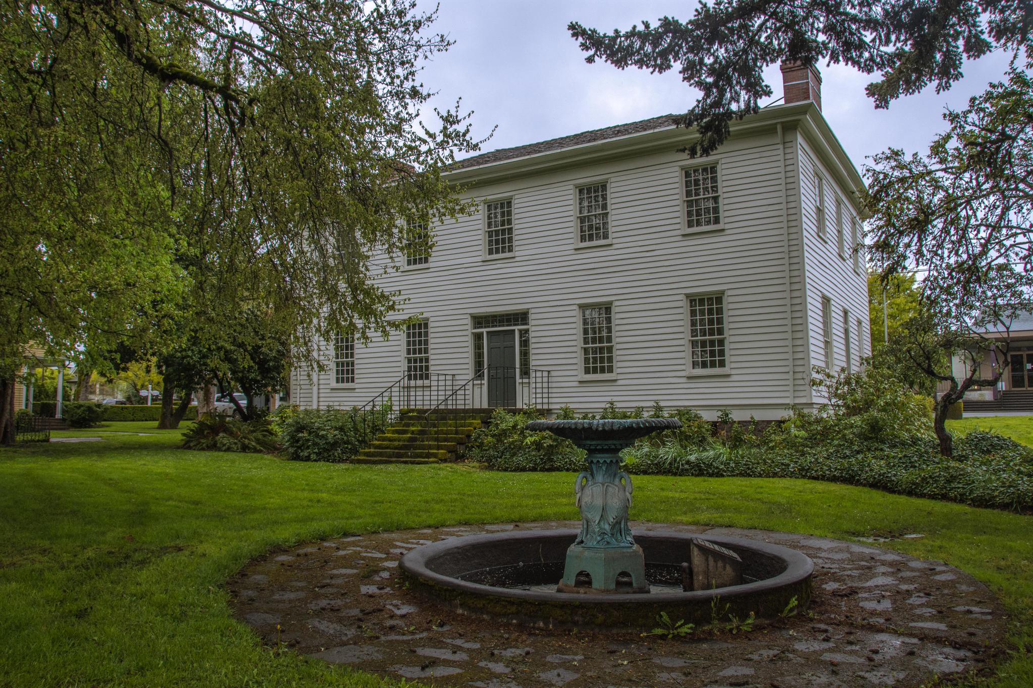 A white historic house with a fountain on a grassy lawn under an overcast sky.
