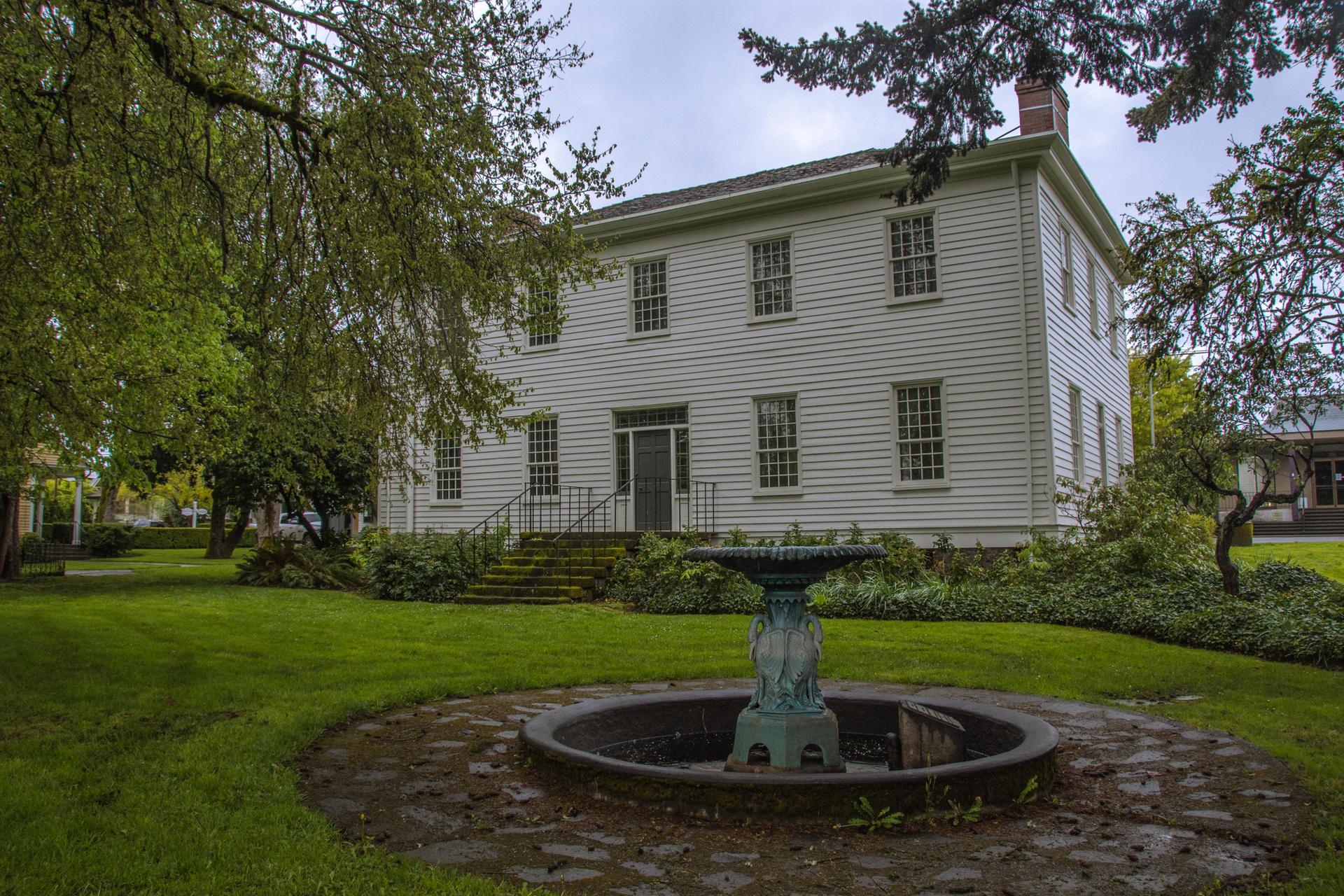 A white historic house with a fountain on a grassy lawn under an overcast sky.