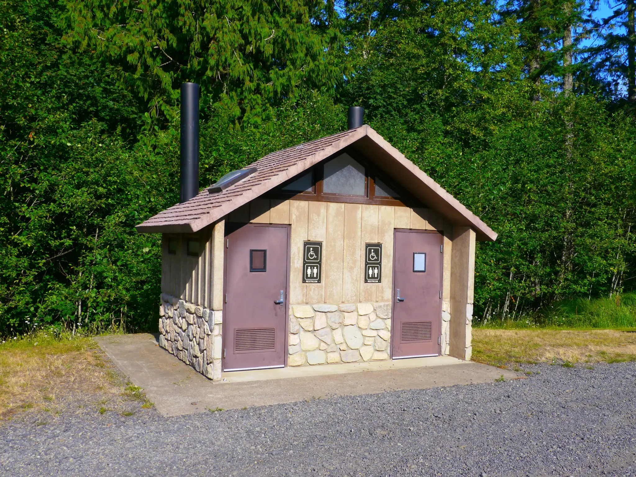 Rustic public restroom building with two doors, surrounded by green trees, bright sun. - Clayhill Horse Staging Area – Hilltop Shelter & Viewpoint loop from L.L. Stub Stewart State Park