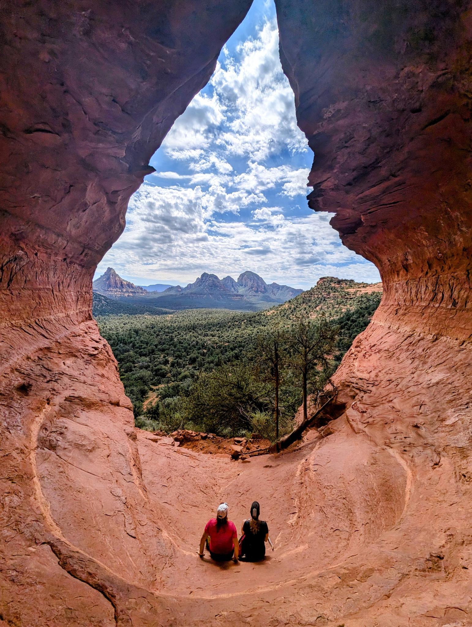 Two people sit in a red rock cave, overlooking a green valley and mountains.