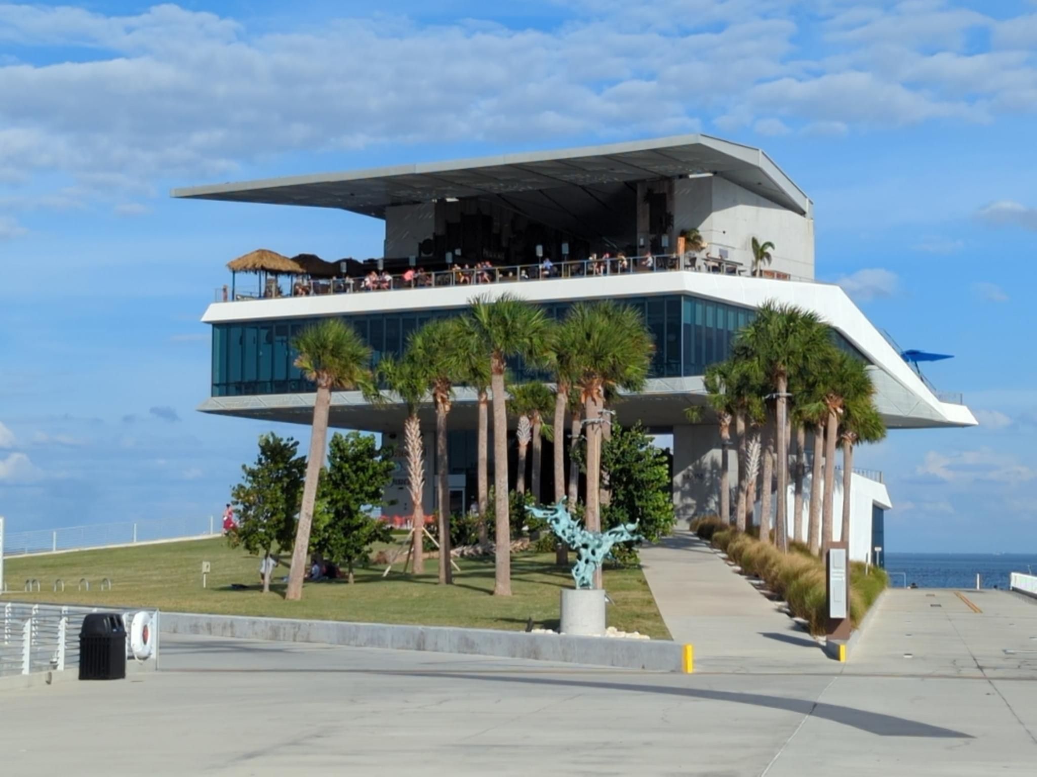 Modern multi-story building with people on deck, surrounded by palm trees. - St. Pete Pier