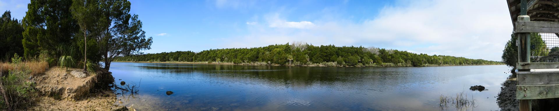 Calm water reflecting a forested shoreline under a bright, partly cloudy sky.