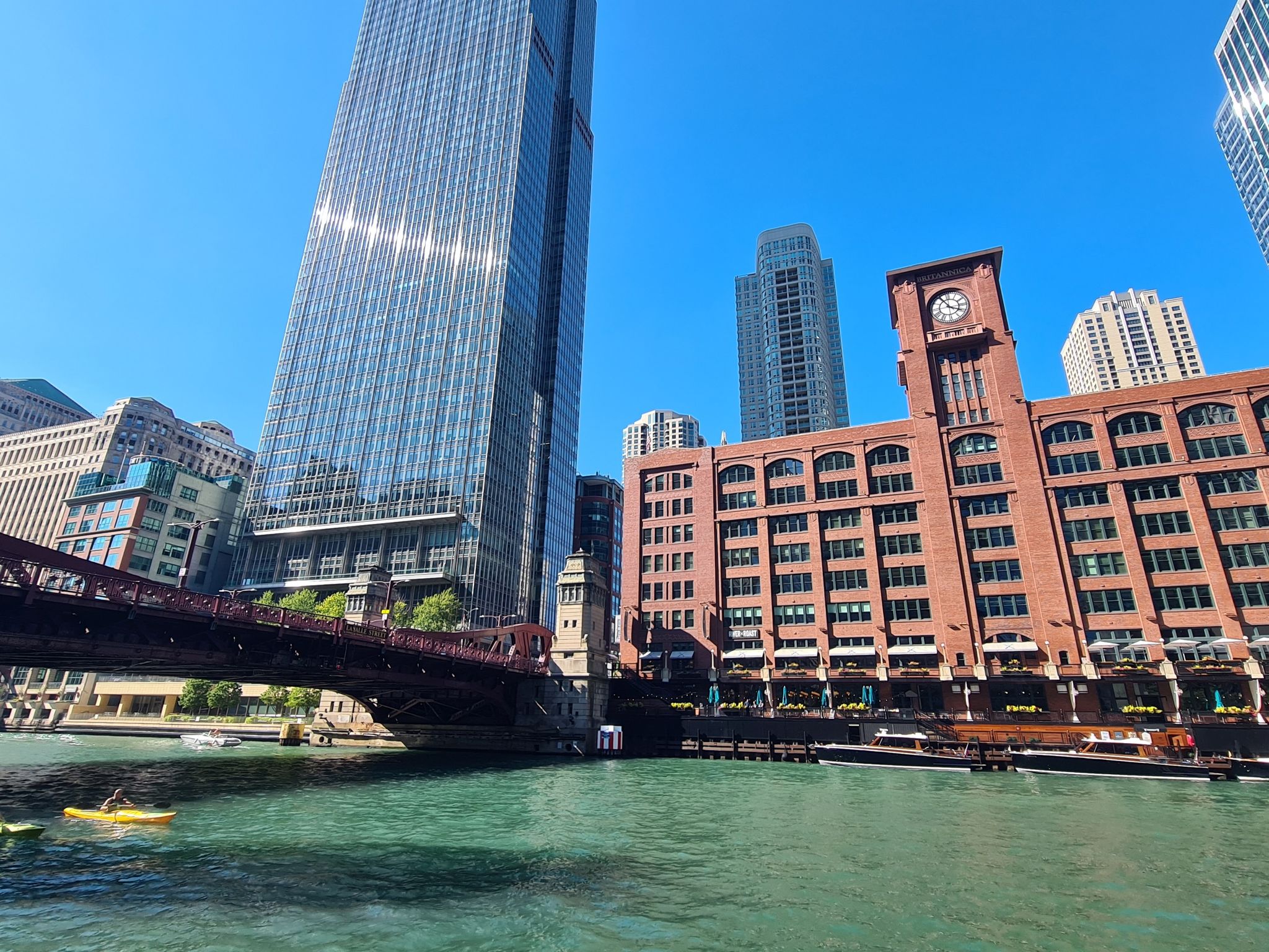 Kayakers paddle on a river surrounded by tall city buildings on a sunny day. - Chicago Riverwalk