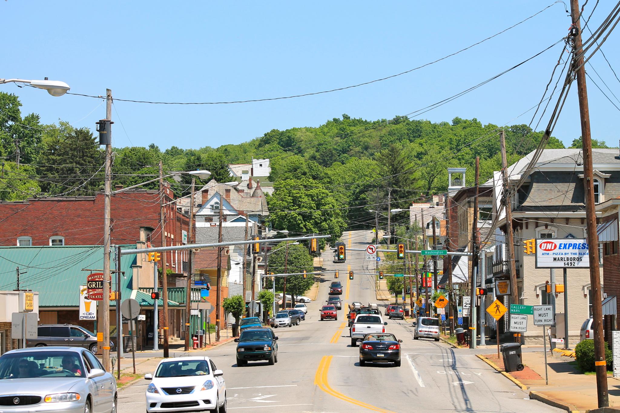 A busy street with cars and buildings, leading uphill under bright sun.