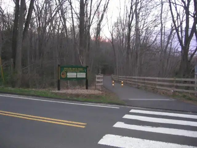 A paved road and crosswalk lead to a wooded path with a sign and fence.