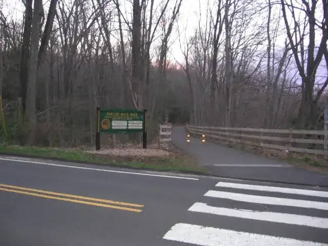A paved road and crosswalk lead to a wooded path with a sign and fence. - Pequonnock Valley Greenway Rails to Trails