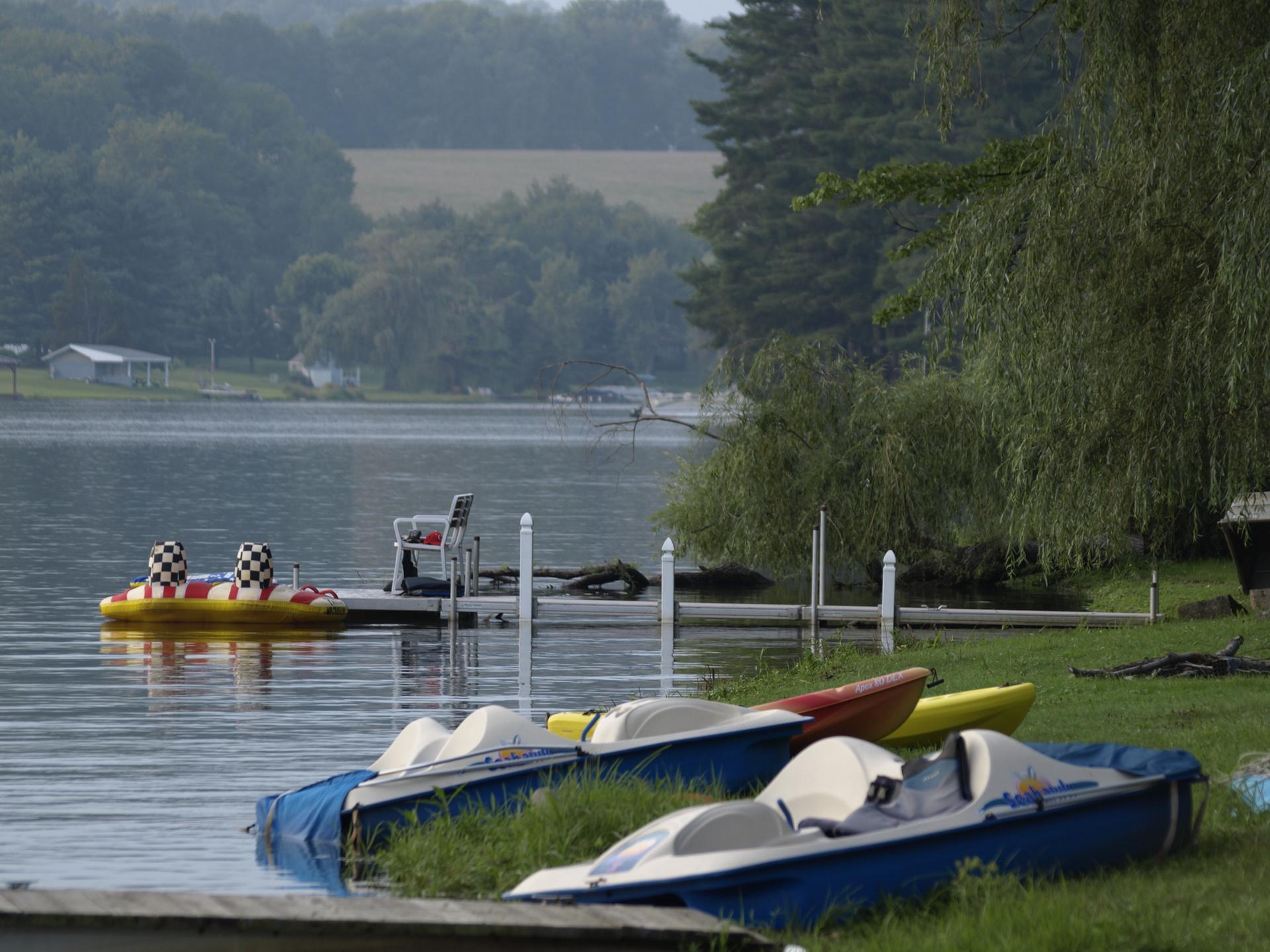 Pedal boats and kayaks rest on a grassy lake shore under soft light.