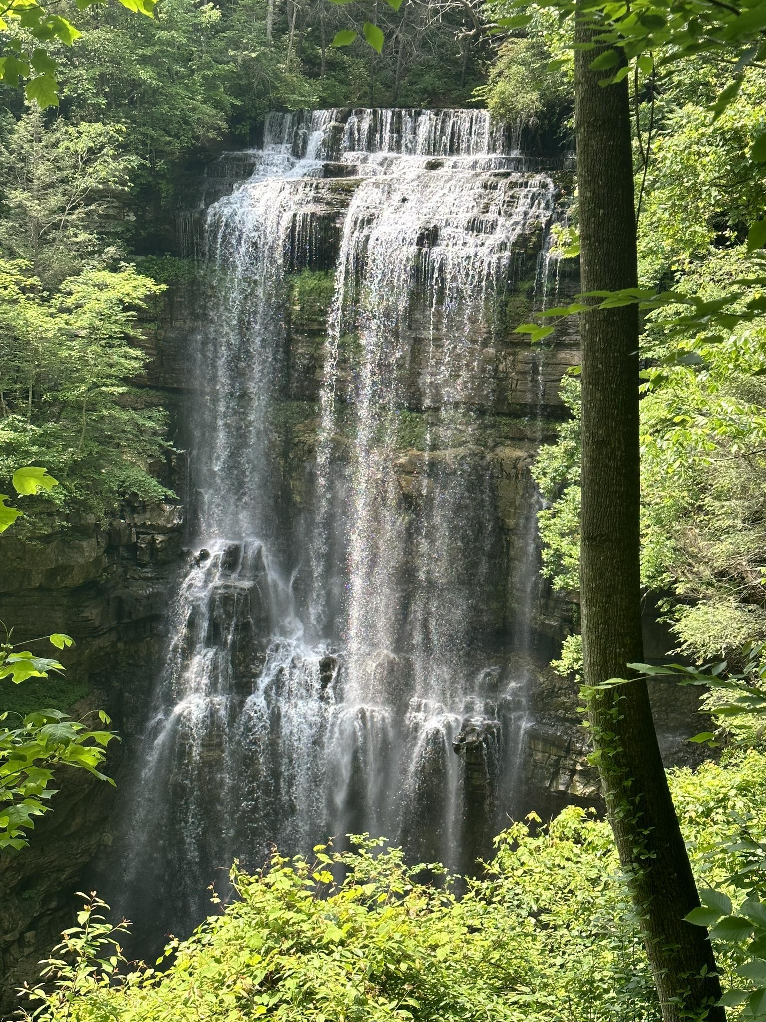 A multi-tiered waterfall cascades down rocky cliffs surrounded by lush green forest.