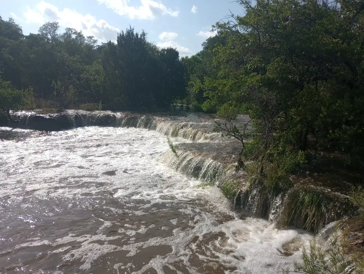 Rushing brown water cascades over a wide, multi-tiered waterfall surrounded by green trees.