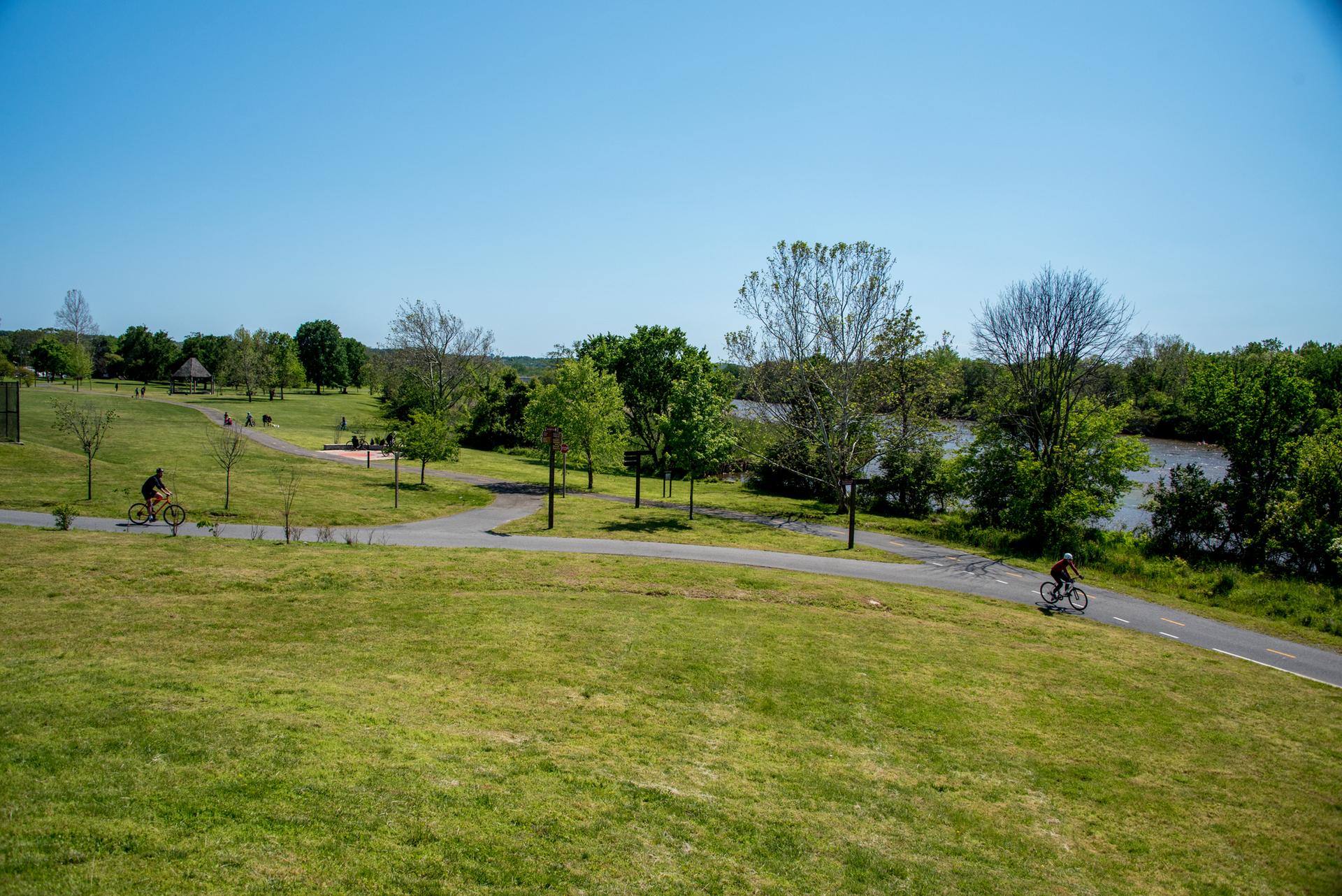 Two people cycling on paved paths through a sunny green park with a river.