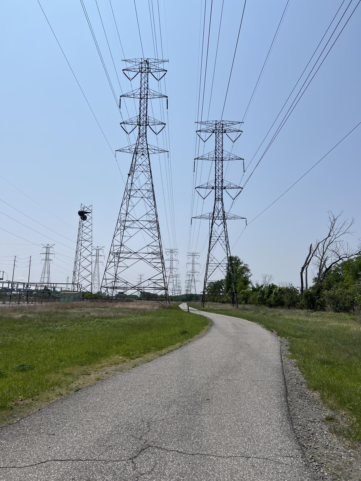 Paved path leading towards large power line towers on a sunny day.